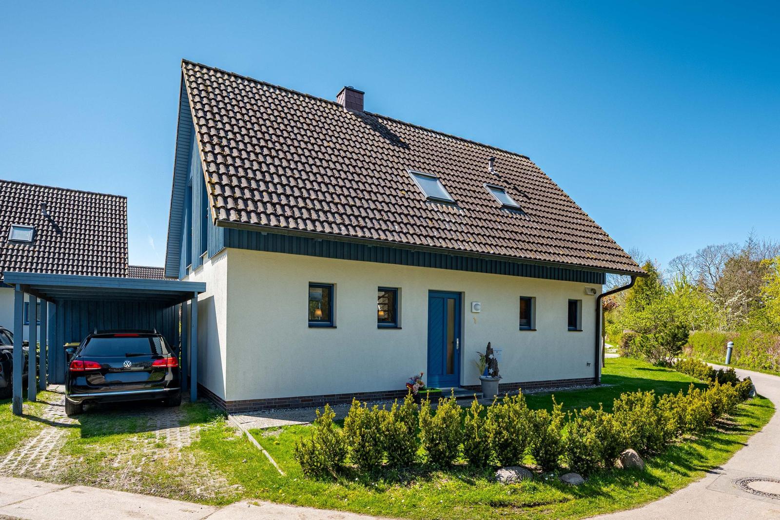 Ein Haus mit Schieferdach, blauer Tür und Garagenbereich. Grünflächen und Bäume im Hintergrund.