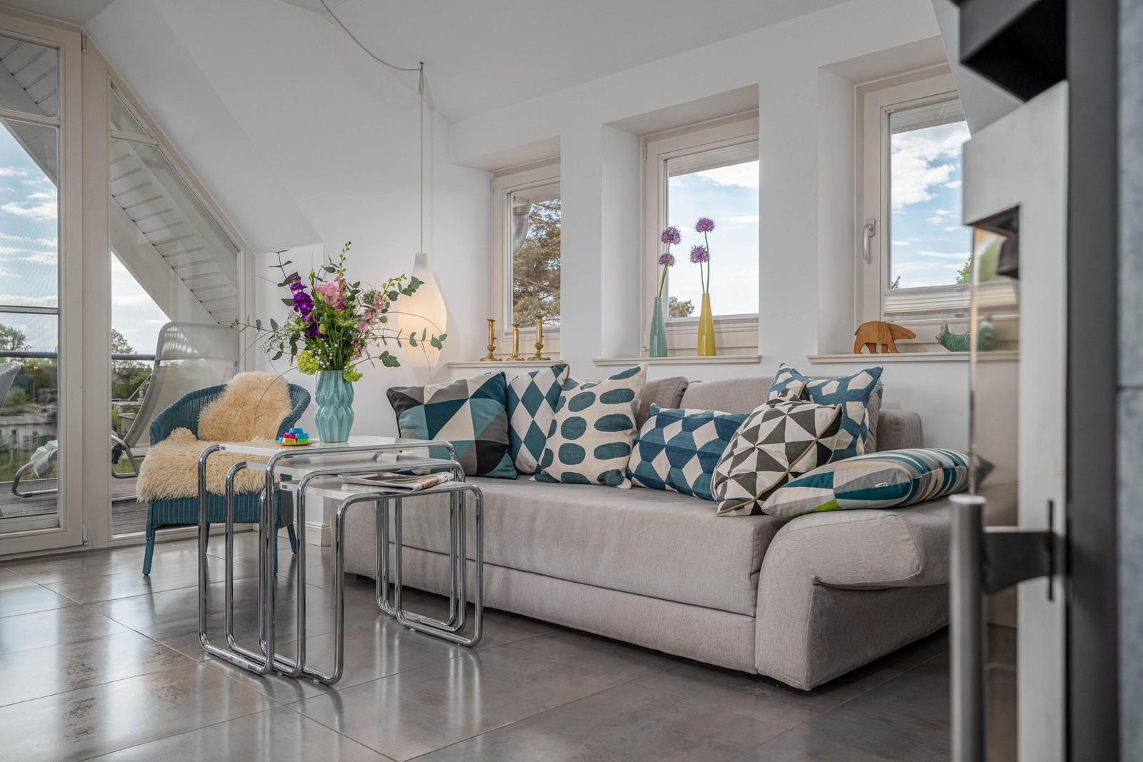 Wohnzimmer mit Sofa, Kissen, Tisch und Fenstern. Draußen ist eine Terrasse.