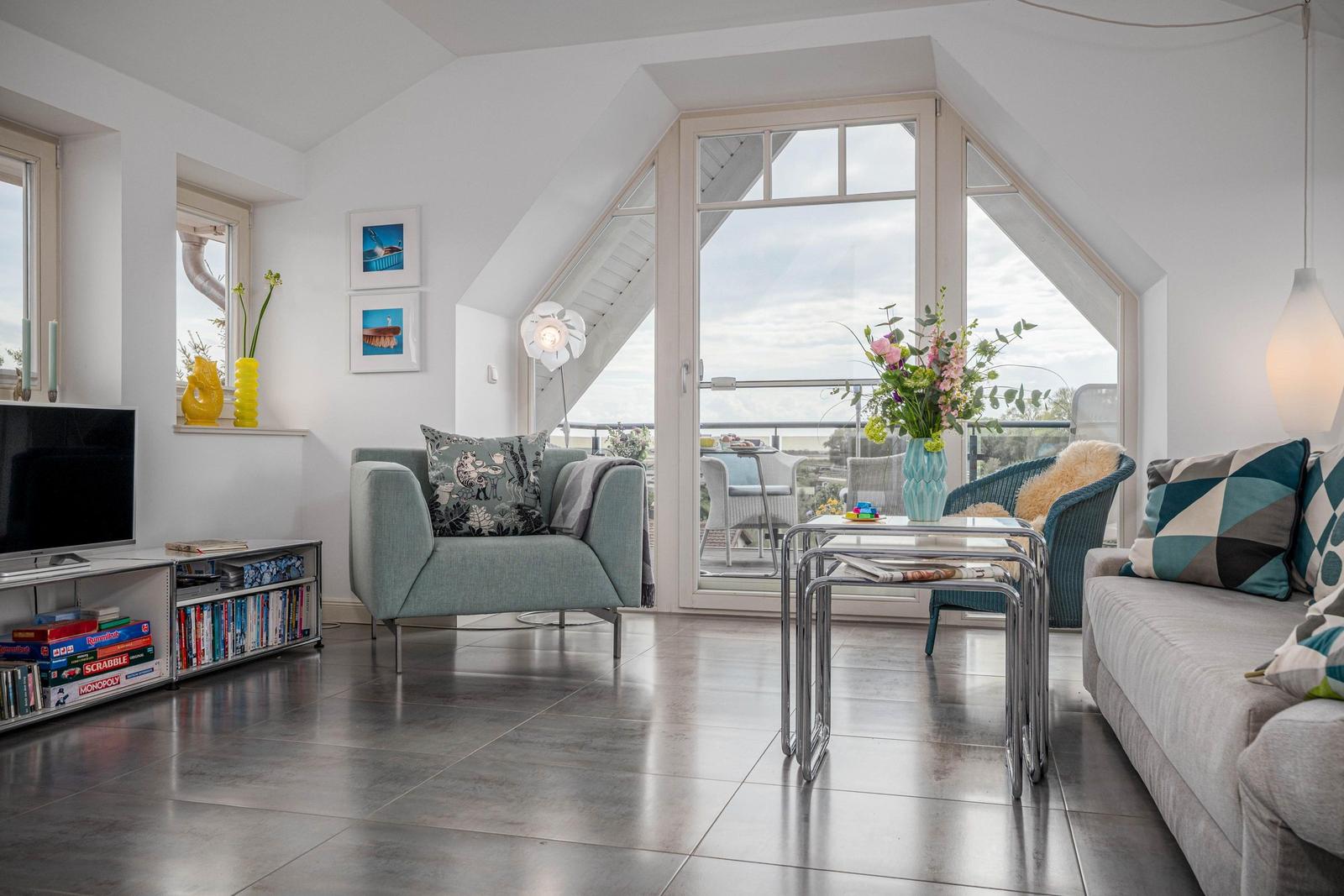Wohnzimmer mit Sitzgelegenheiten, TV und Balkonzugang. Fliesenboden und große Fenster.