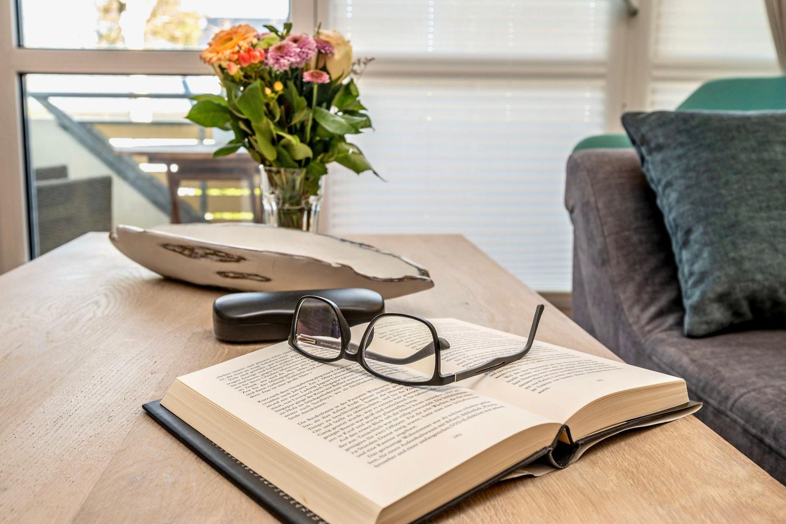 A table with a book, glasses, and flowers by a window.