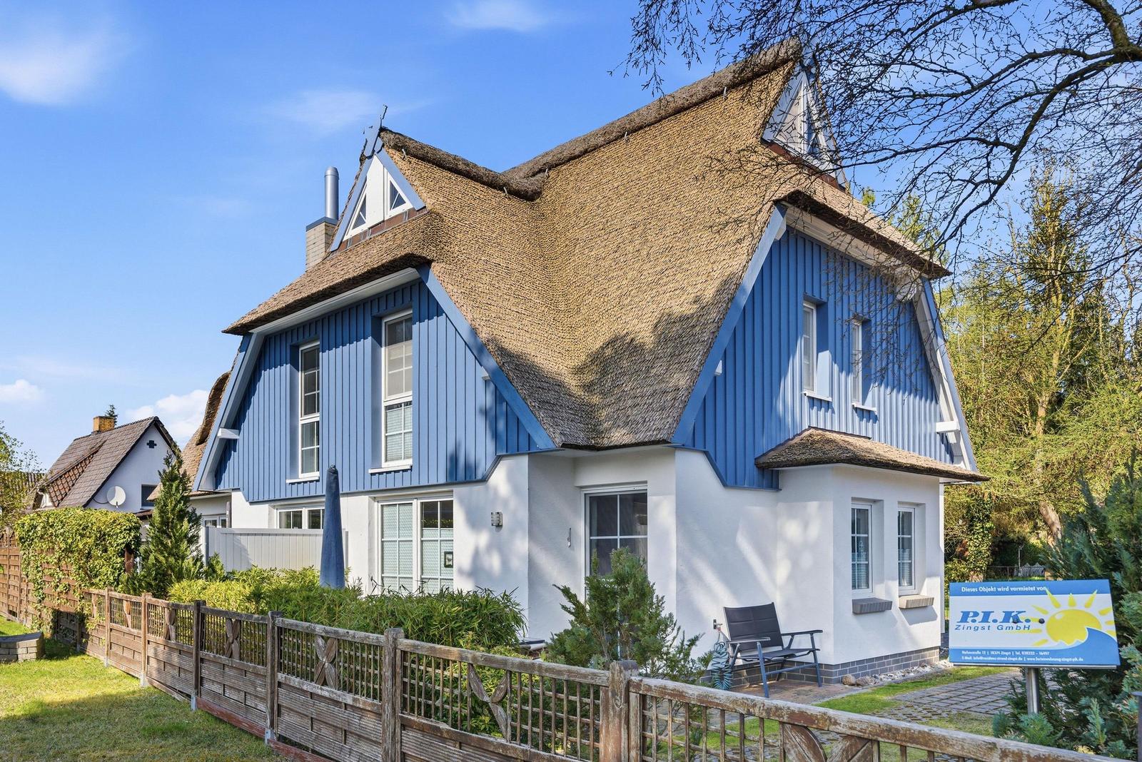 Ein zweistöckiges Haus mit blauen und weißen Wänden und strohgedecktem Dach. Garten mit Holzzaun.