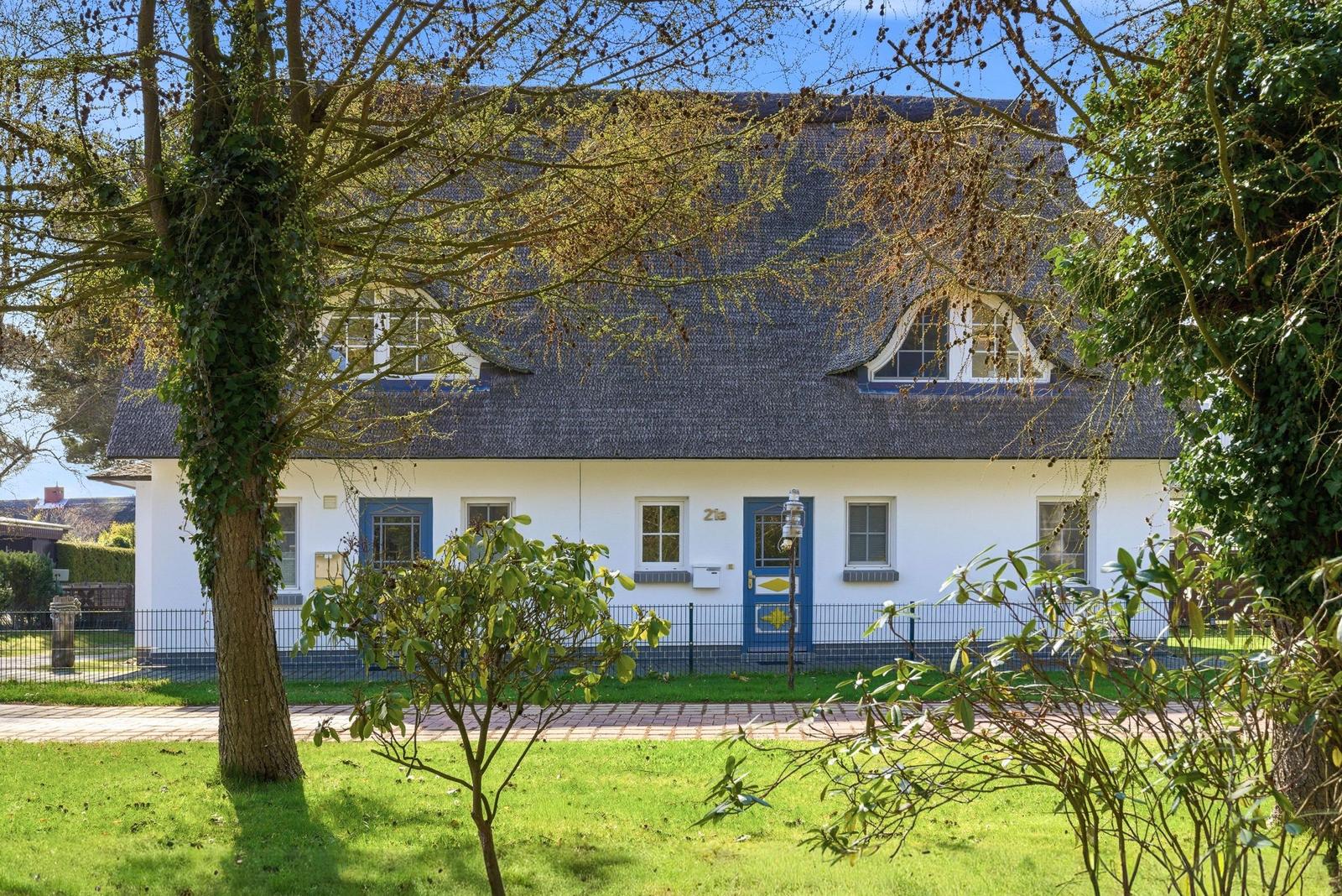 Ein weißes Haus mit blauen Fenstern und Tür, Dach aus Schindeln, umgeben von Grünflächen und Bäumen.