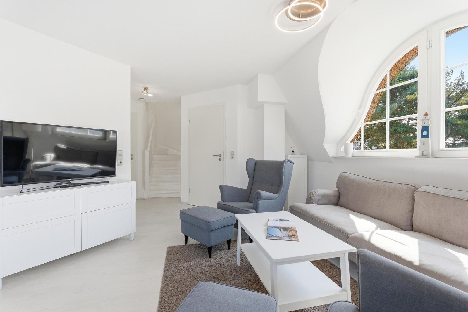 Wohnzimmer mit Sofa, Sessel und Fernseher. Große Fenster mit Blick auf Bäume.