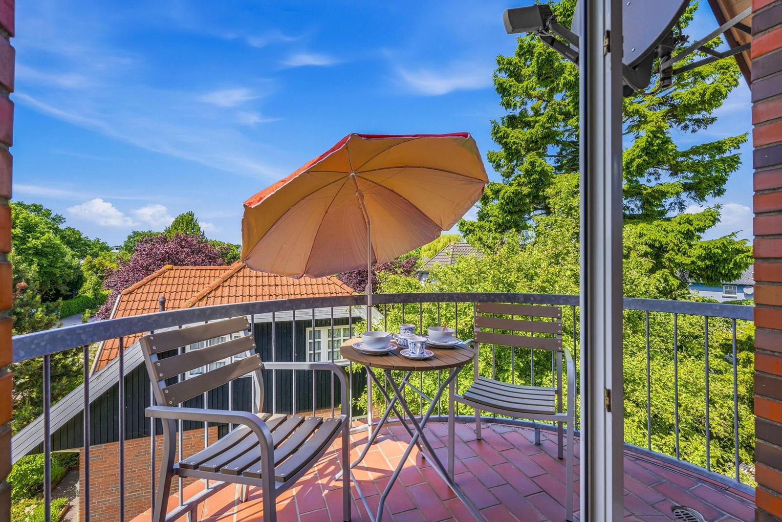 Terrasse mit Tisch, zwei Stühlen und Sonnenschirm. Blick auf grüne Bäume und ein Haus mit rotem Dach.
