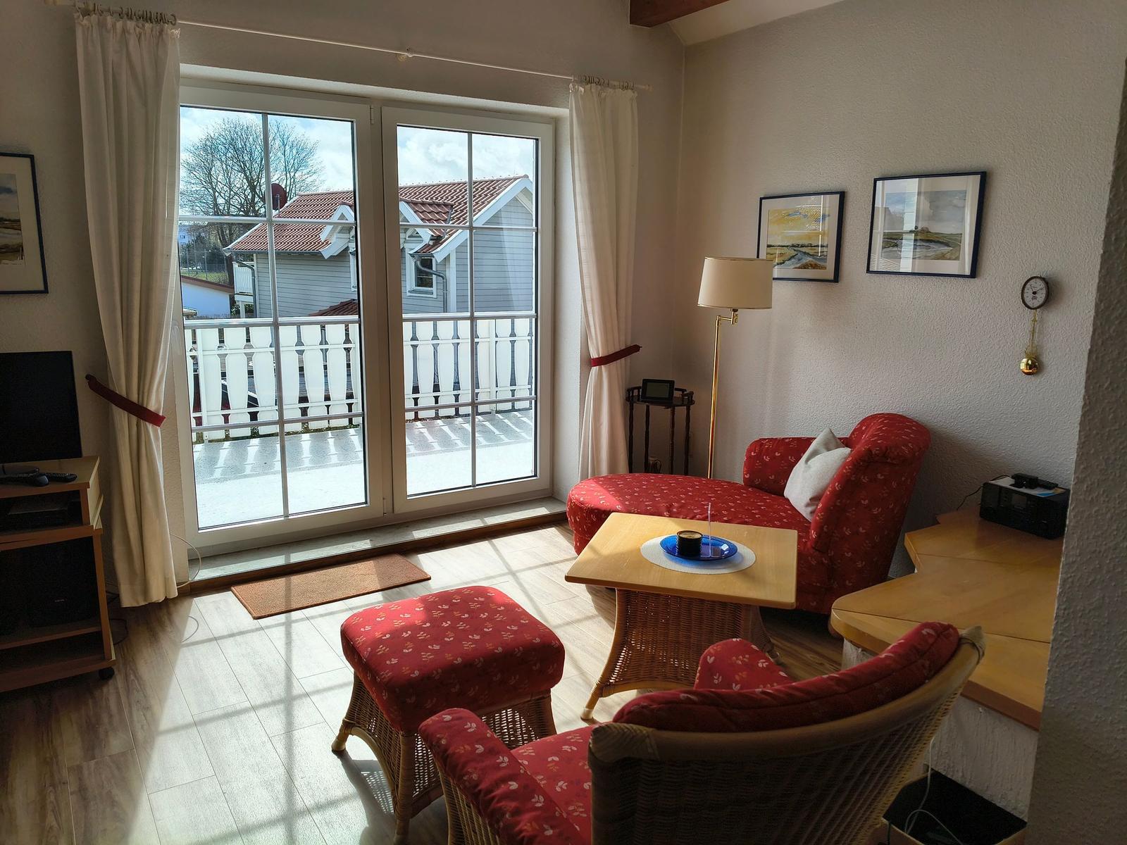 Wohnraum mit rotem Sofa, Sitzgelegenheiten und Balkon. Fenster und Wandbilder sichtbar.