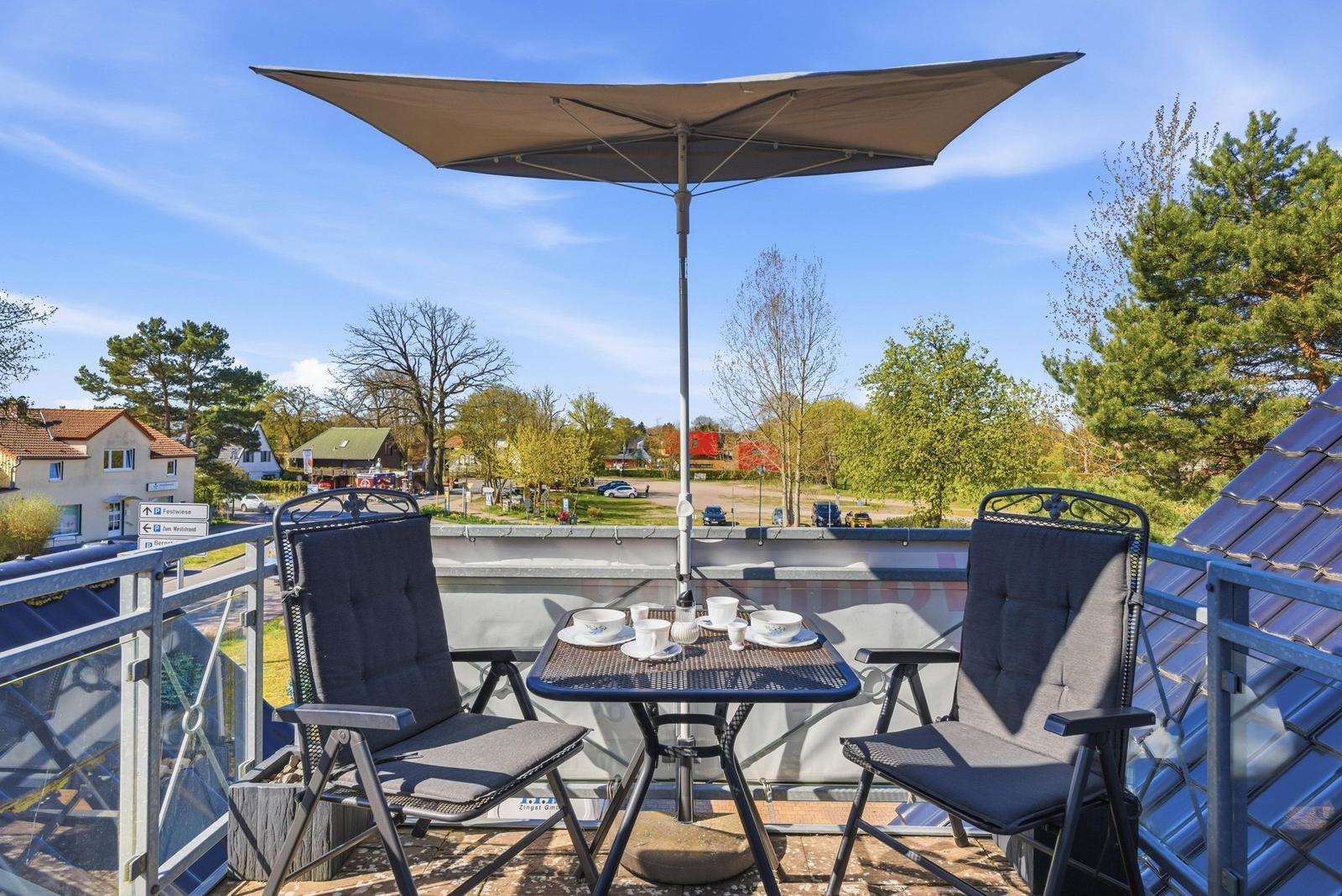 Terrasse mit Tisch, zwei Stühlen und Sonnenschutz. Blick auf Straße und Bäume.