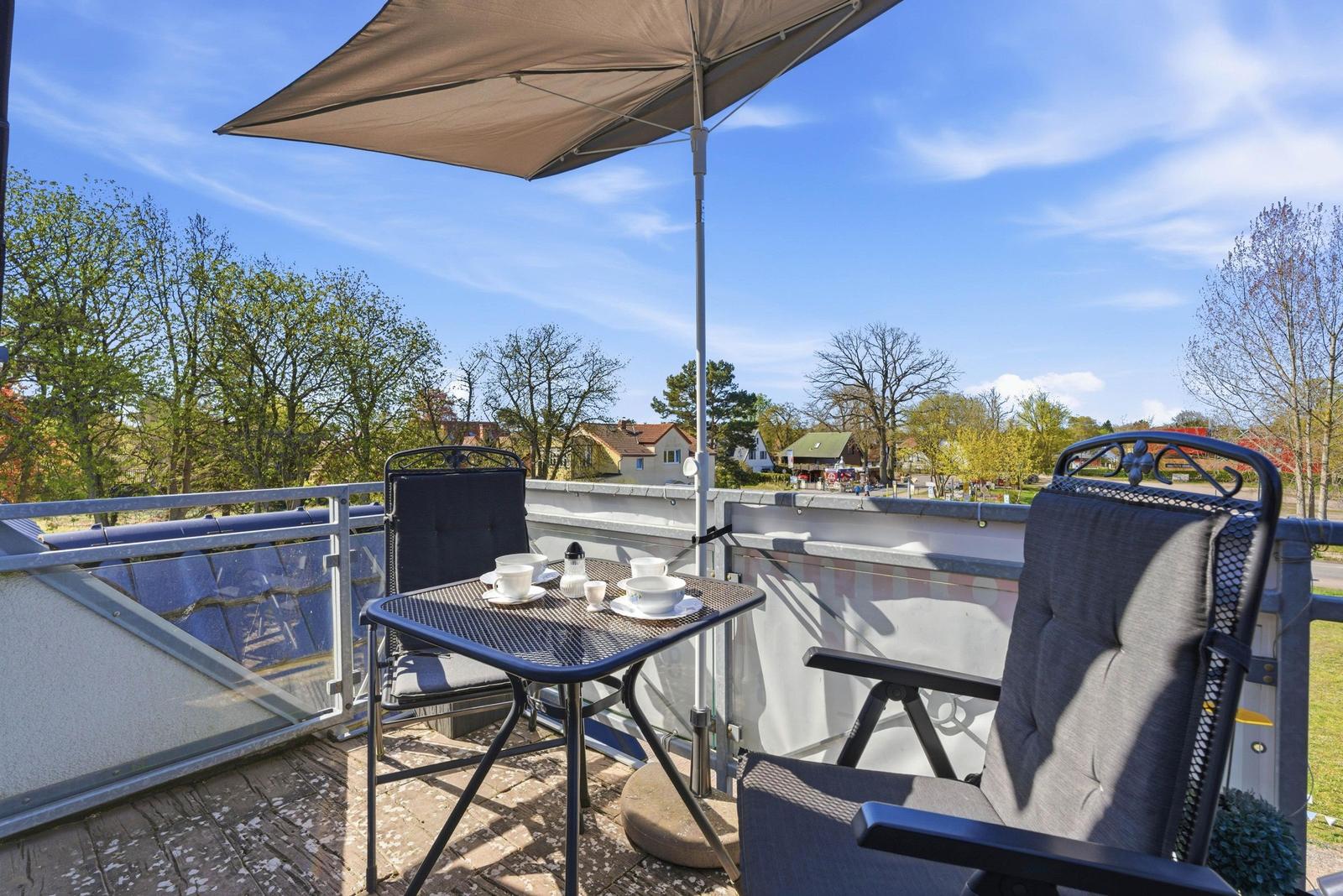 Terrasse mit Tisch, zwei Stühlen und Sonnenschutz. Blick auf Bäume und Häuser.