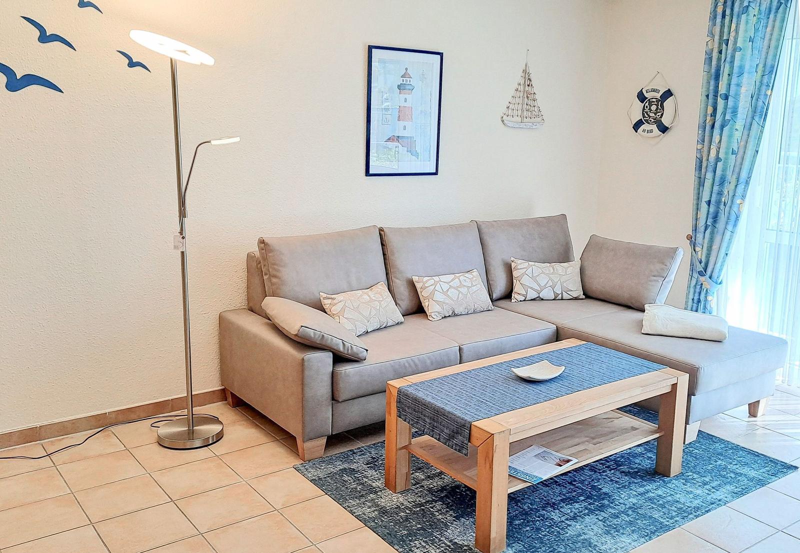 Ein Wohnzimmer mit grauem Sofa, Holz-Tisch und blauem Teppich. Wanddekoration und Vorhänge im Seemannsstil.