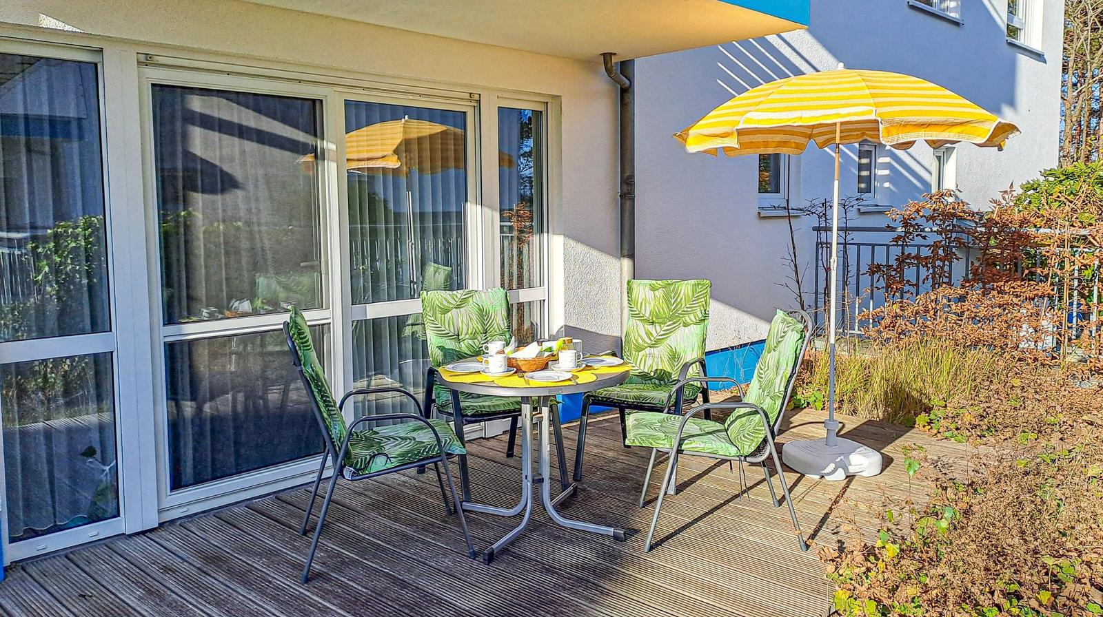Terrasse mit Tisch, vier Stühlen und gelbem Sonnenschirm. Holzdielen und Pflanzen umgeben den Außenbereich.