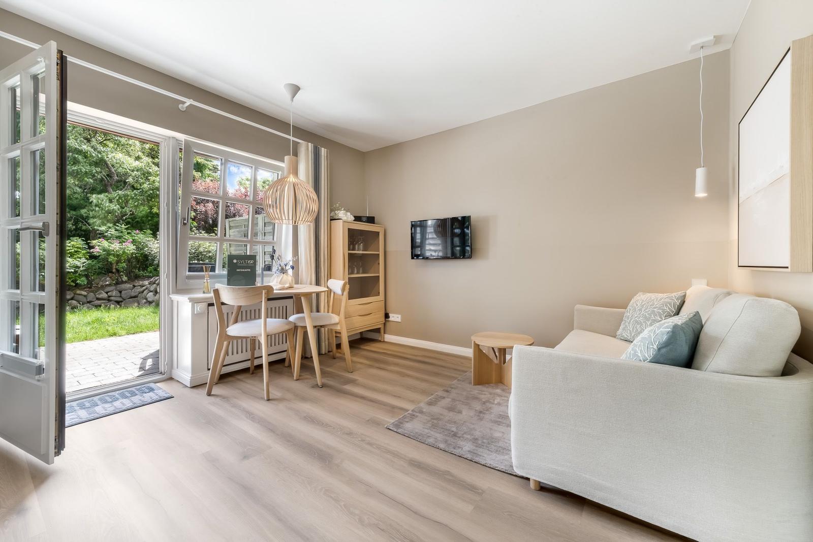 Wohn- und Essbereich mit Sofa, Tisch, Stühlen und TV. Durchgang zur Terrasse mit Gartenblick.
