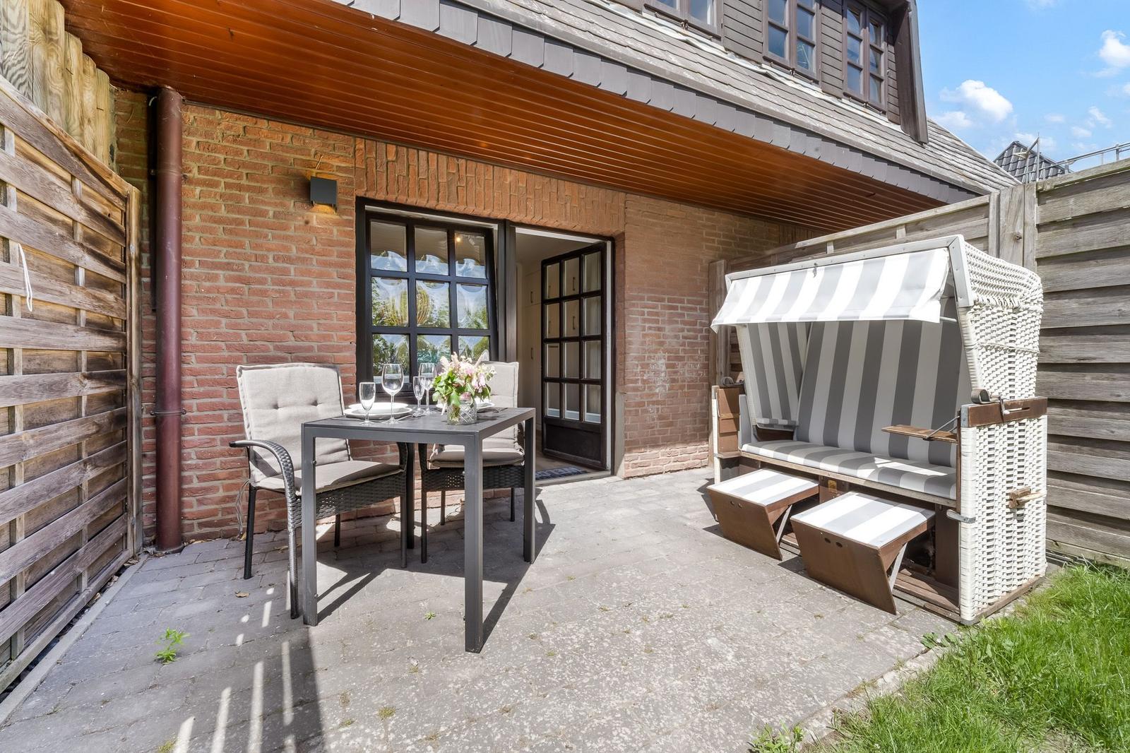 Terrasse mit Tisch, Stühlen und Strandkorb. Hintergrund: Backsteinhaus mit Fenstern.