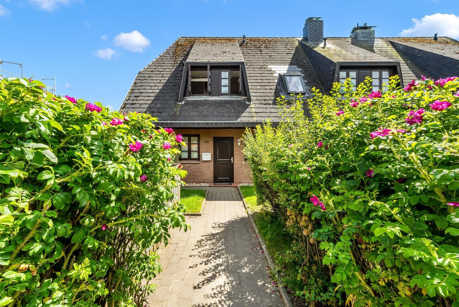 Haus mit schwarzem Dach, Blumenhecken und gepflastertem Weg zur Tür.