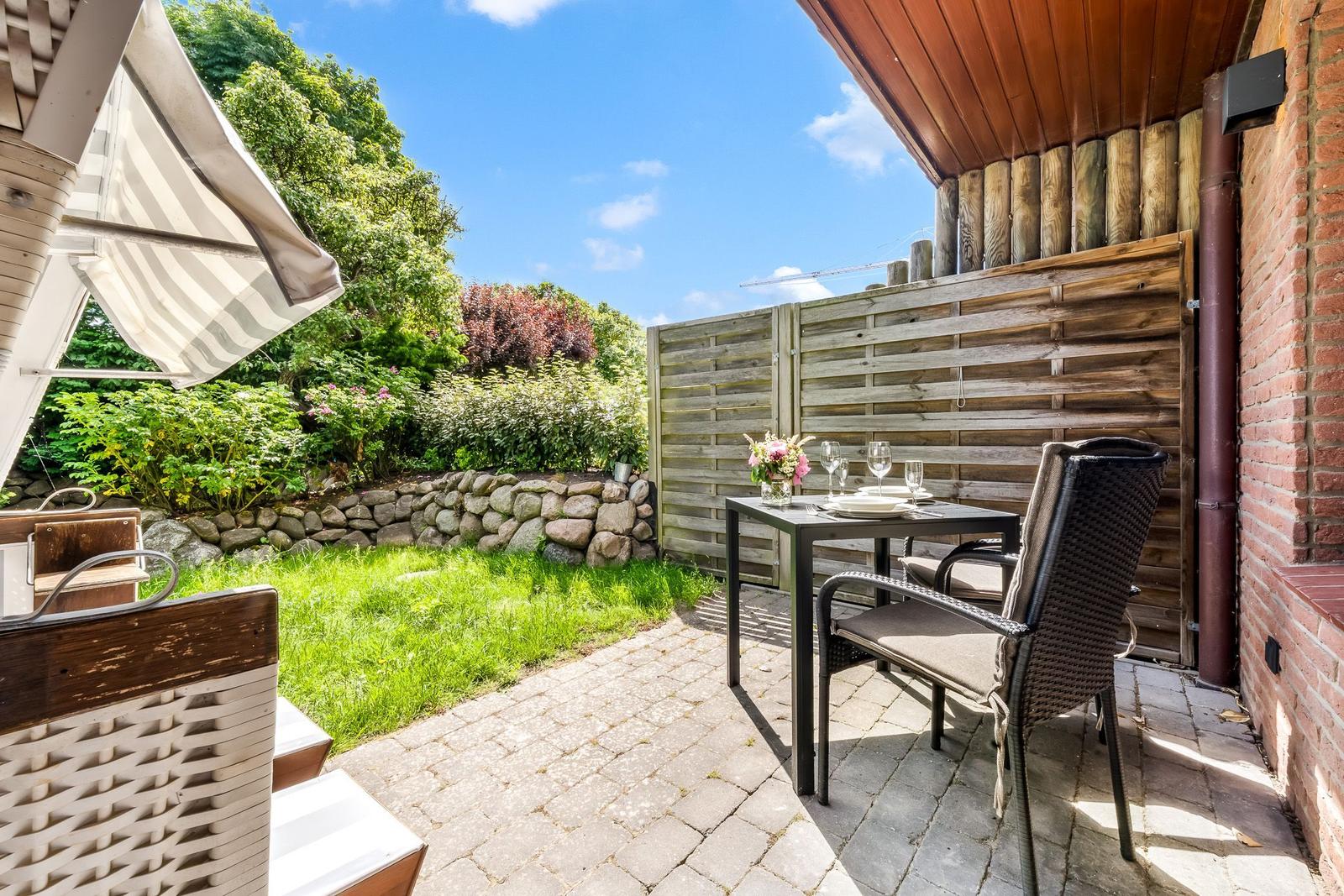 Terrasse mit Tisch, Stühlen und Sonnenschutz. Hintergrund: Grünfläche, Steinmauer und Holzzaun.