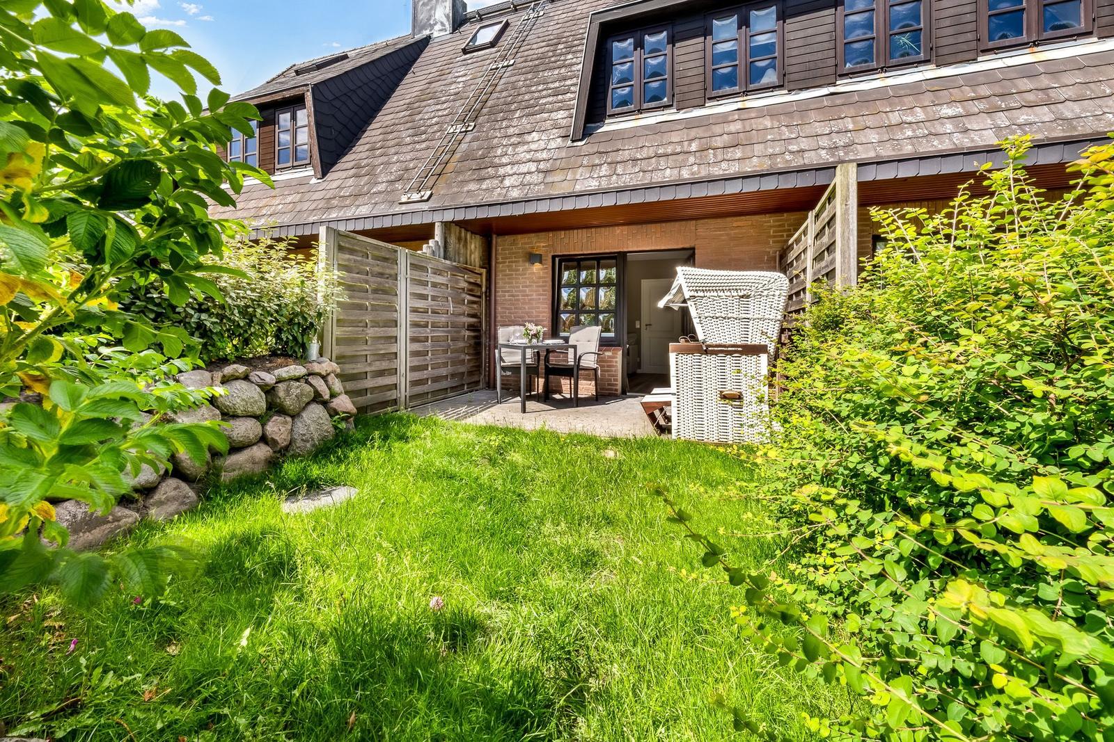 Haus mit Garten, Terrasse und Gartenmöbeln. Holzverkleidung und Steinmauer sichtbar.