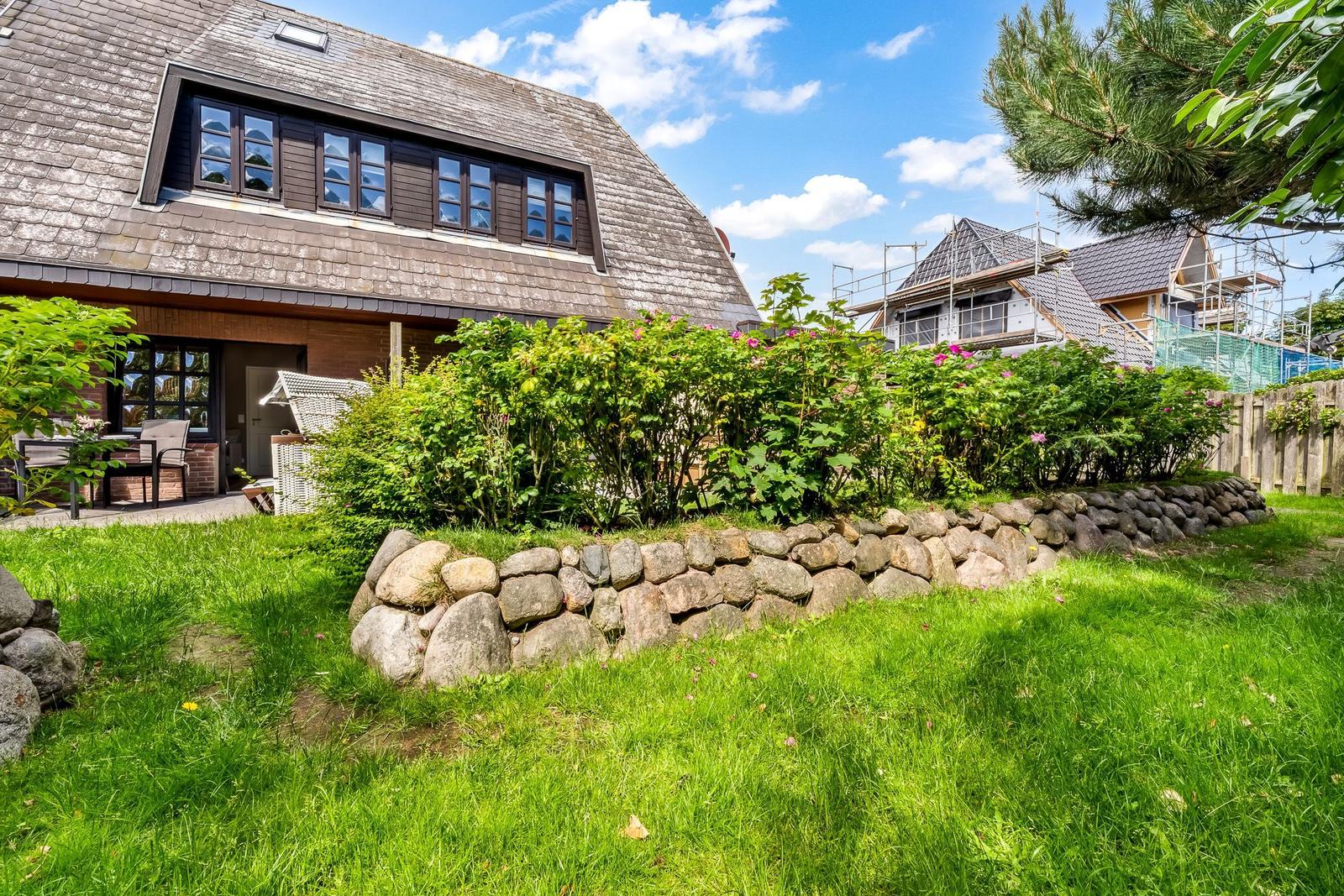 Haus mit Steinmauer, grünem Garten und Blumenhecke unter blauem Himmel.