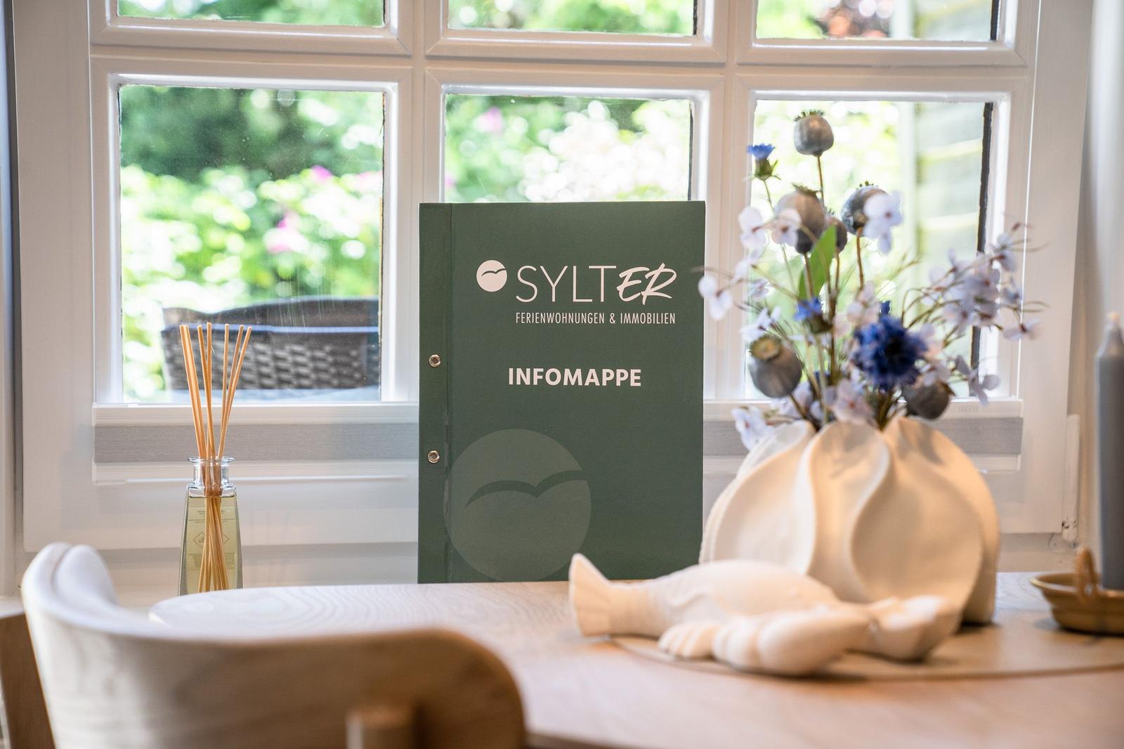 Tisch mit Informationsmappe, Blumen und Duftkerze vor Fenster mit Blick auf Garten.