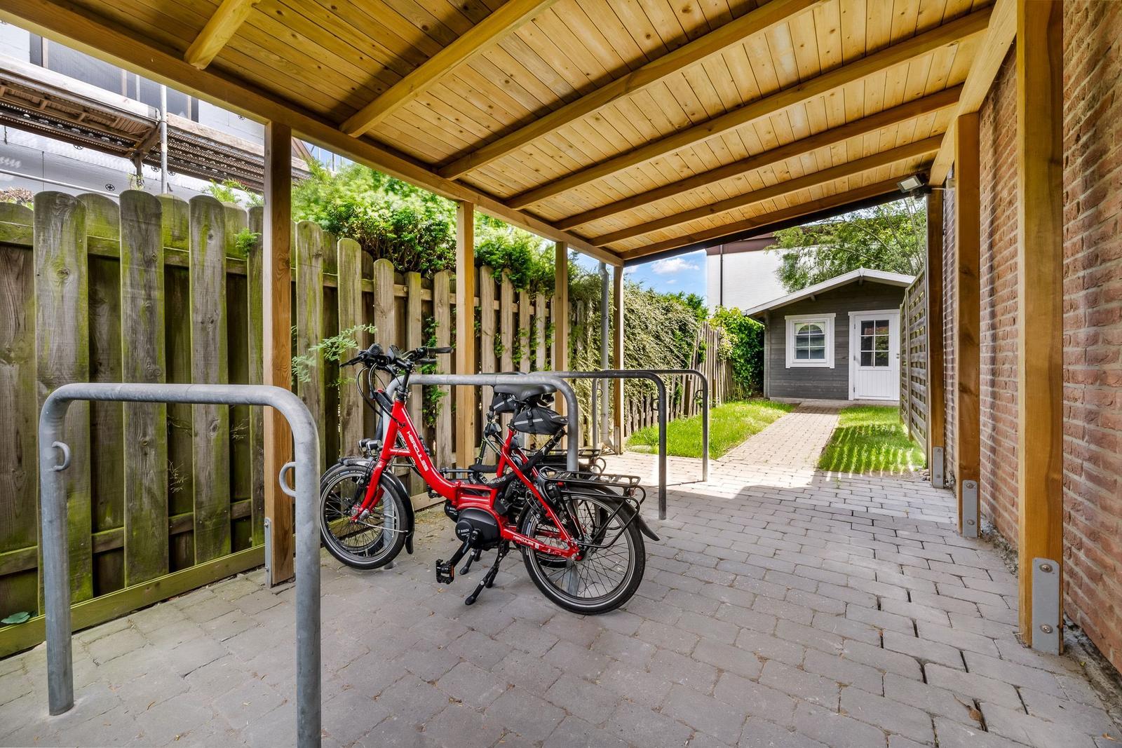 Roter E-Bike unter Holzüberdachung mit Pflasterweg und Gartenhaus im Hintergrund.