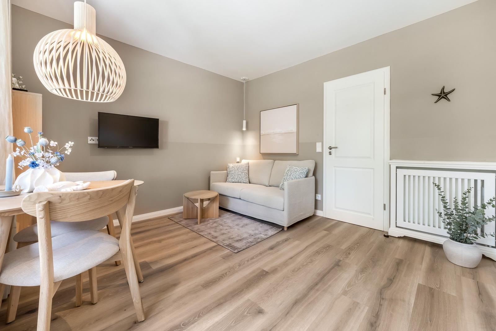 Wohn- und Essbereich mit Sofa, TV und Holzmöbeln. Lichtdurchfluteter Raum mit Holzboden.