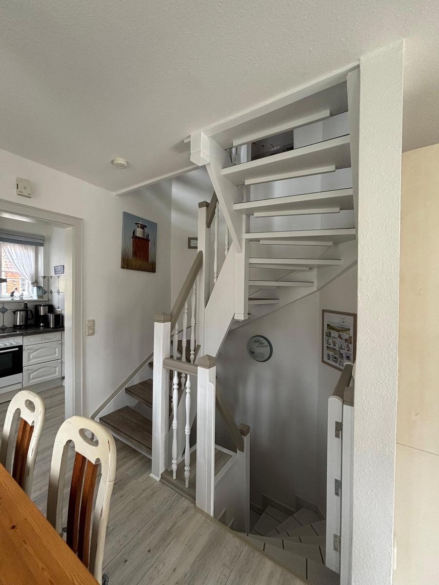 Treppenhaus mit Holzstufen und weißer Balustrade. Küche und Essbereich sichtbar.