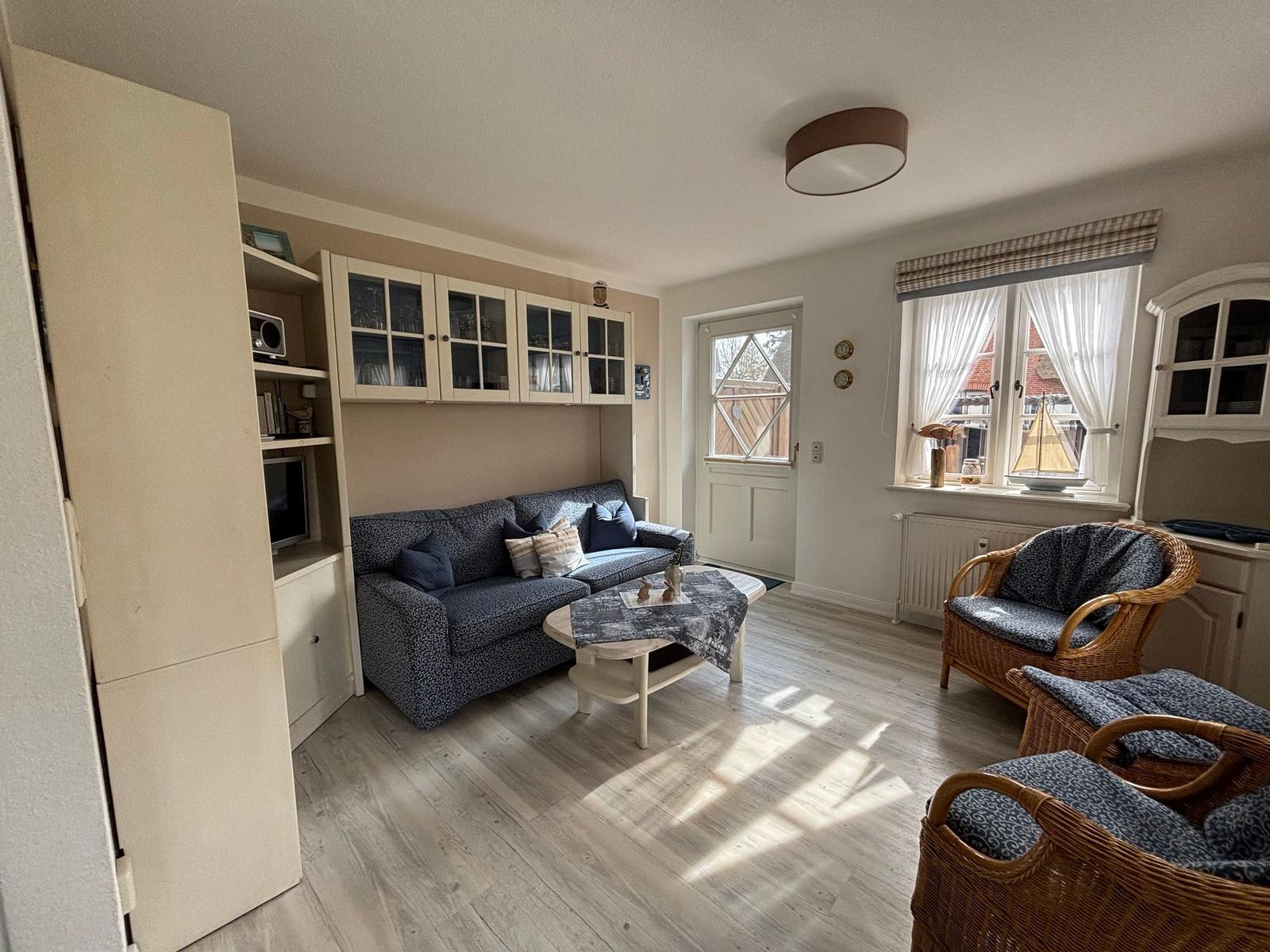 Wohnzimmer mit Sofa, Rattanstühlen und Fensterblick. Holzboden und Küchenecke sichtbar.