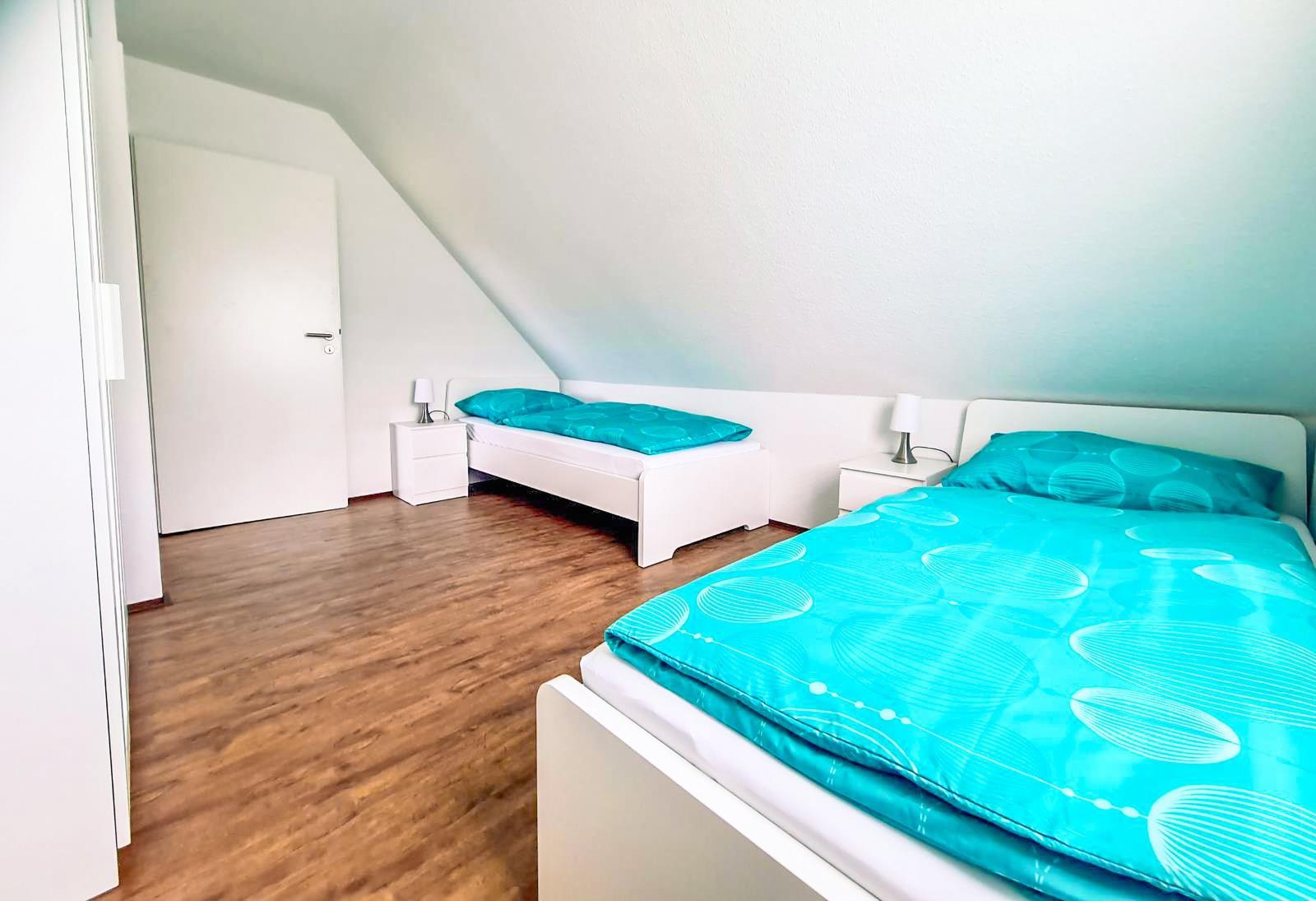 Zwei Betten mit türkisfarbenen Bettwäsche, weiße Nachttische und Holzboden in einem Dachzimmer.