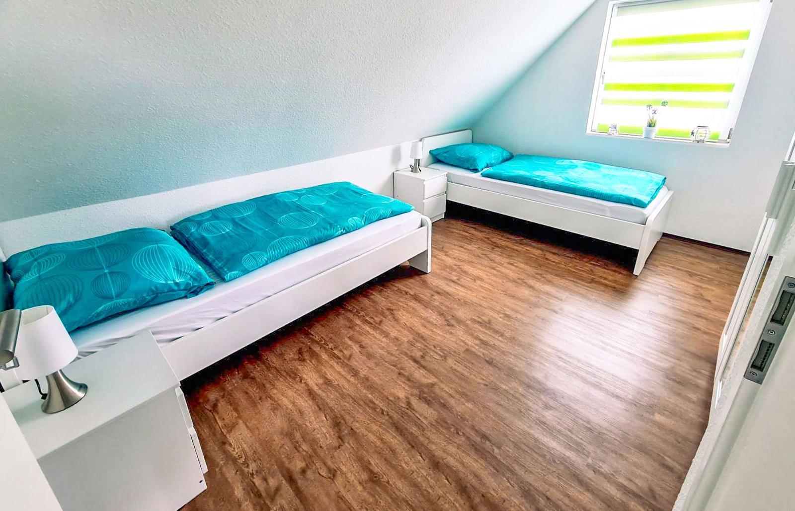 Zimmer mit zwei weißen Betten und Holzboden. Fenster mit grünen Jalousien.