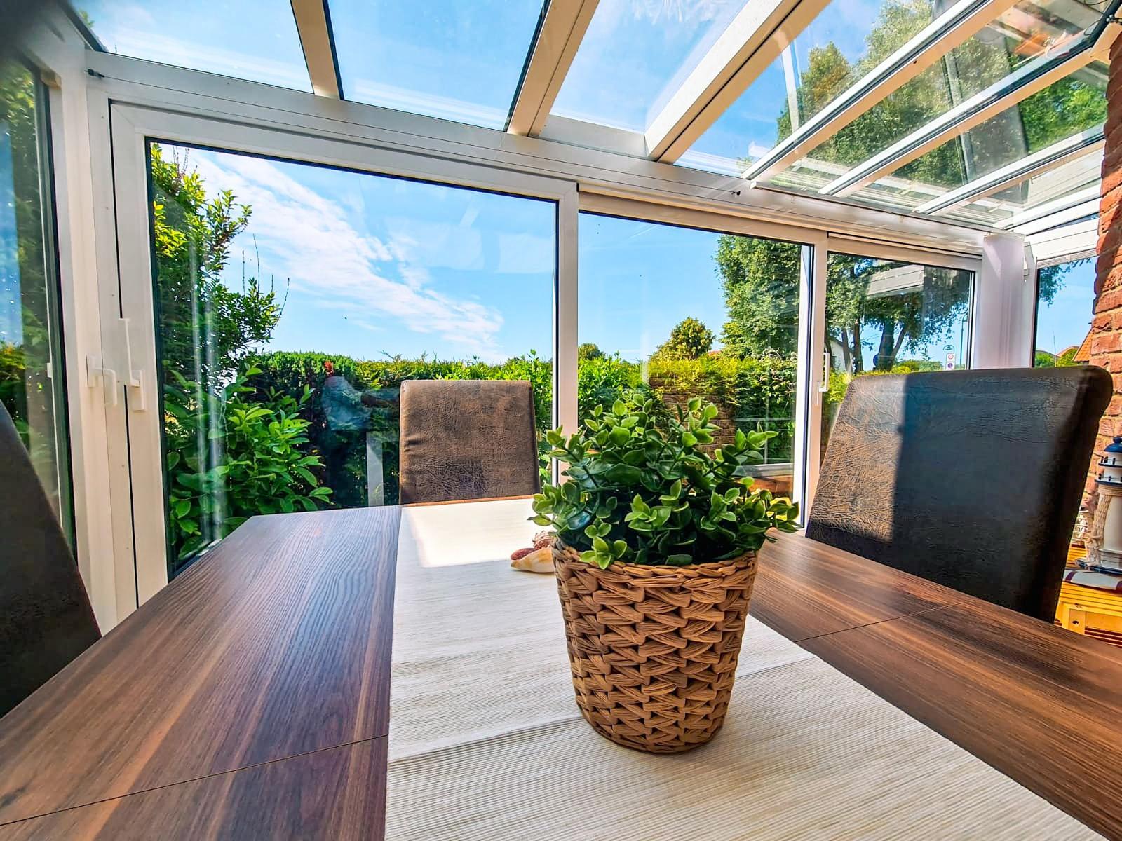 Esstisch mit Stühlen und Pflanze im Glasdachbereich mit Blick auf Garten.