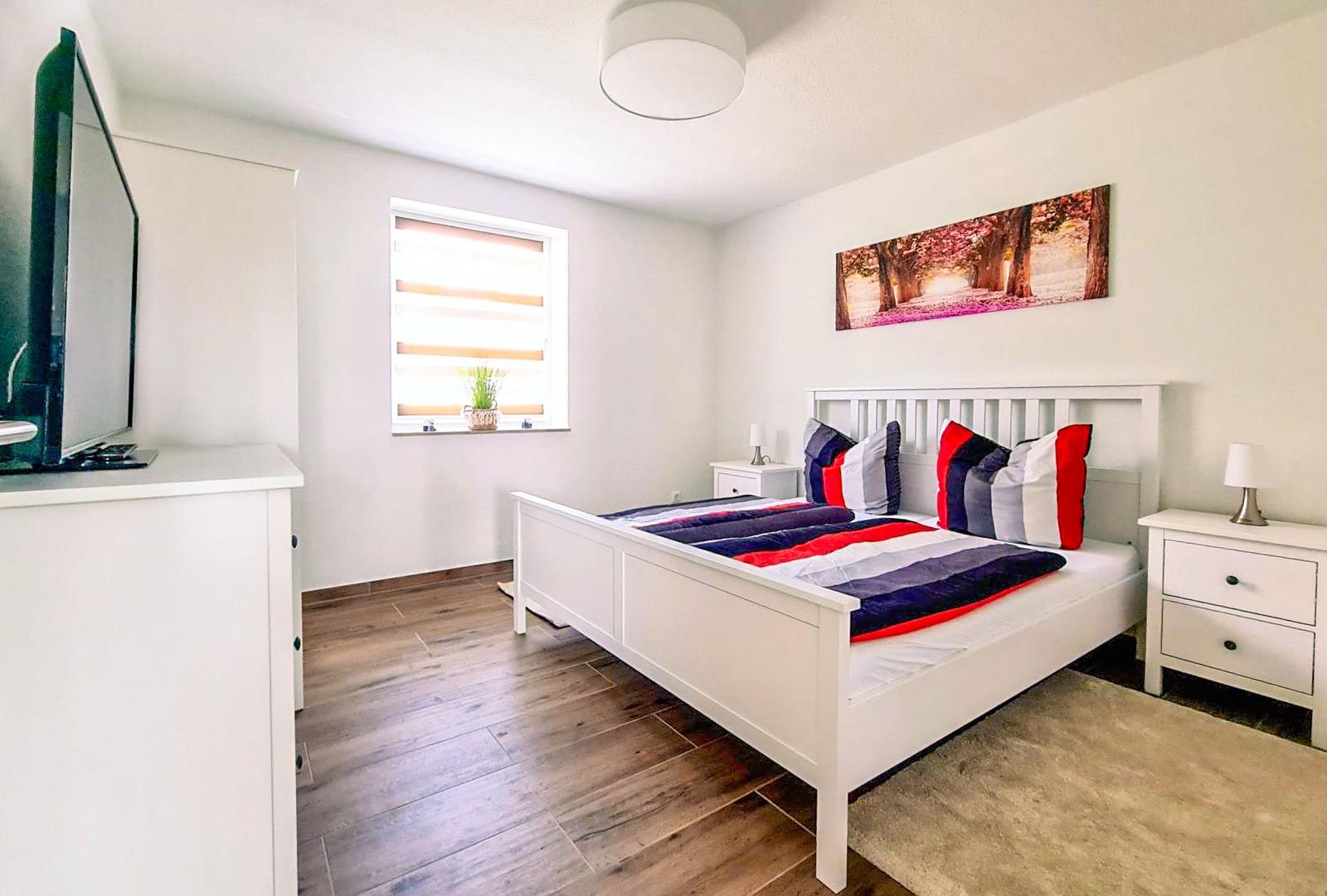 Schlafzimmer mit weißem Bett, TV-Schrank und Fenster mit Jalousie.