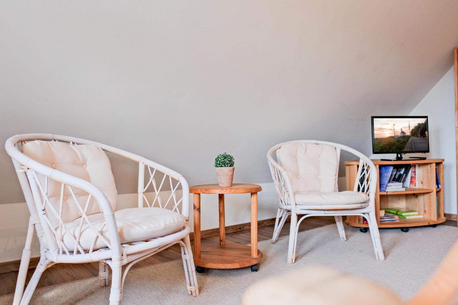 Zwei weiße Rattan-Stühle, kleiner Holztisch mit Pflanze und Fernseher auf Regal.