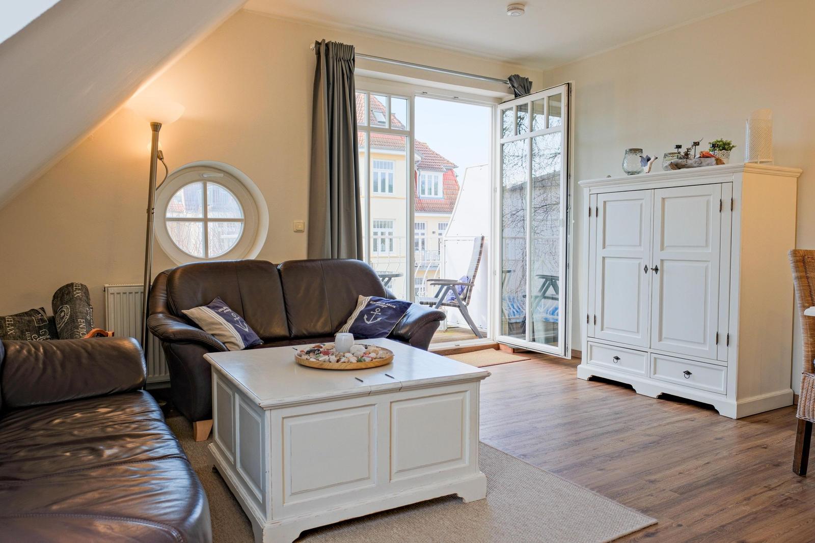 Wohnzimmer mit braunem Sofa, weißem Tisch und Schrank. Fenster mit Blick auf Balkon.