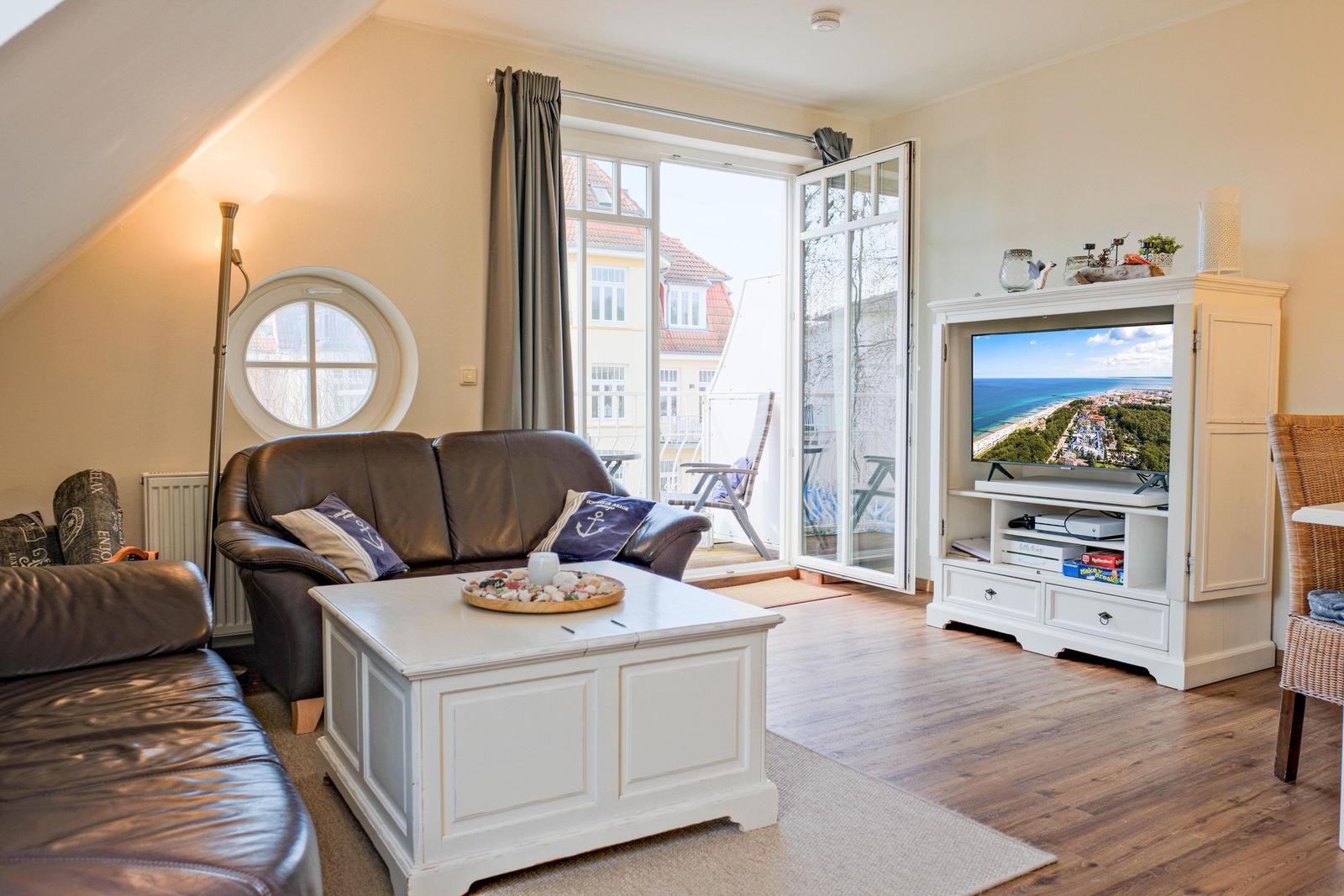 Wohnzimmer mit Sofa, Couchtisch und TV. Balkon mit Sitzgelegenheit. Rundes Fenster. Holzboden.