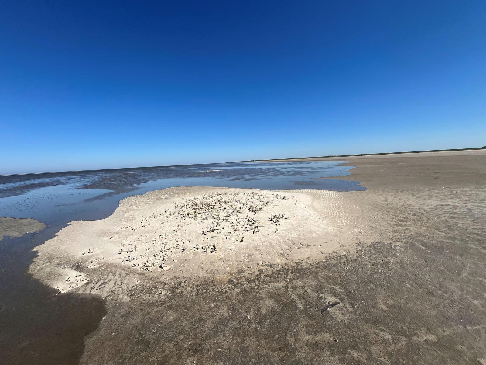 Strand mit flachem Wasser und Sandinsel unter blauem Himmel.