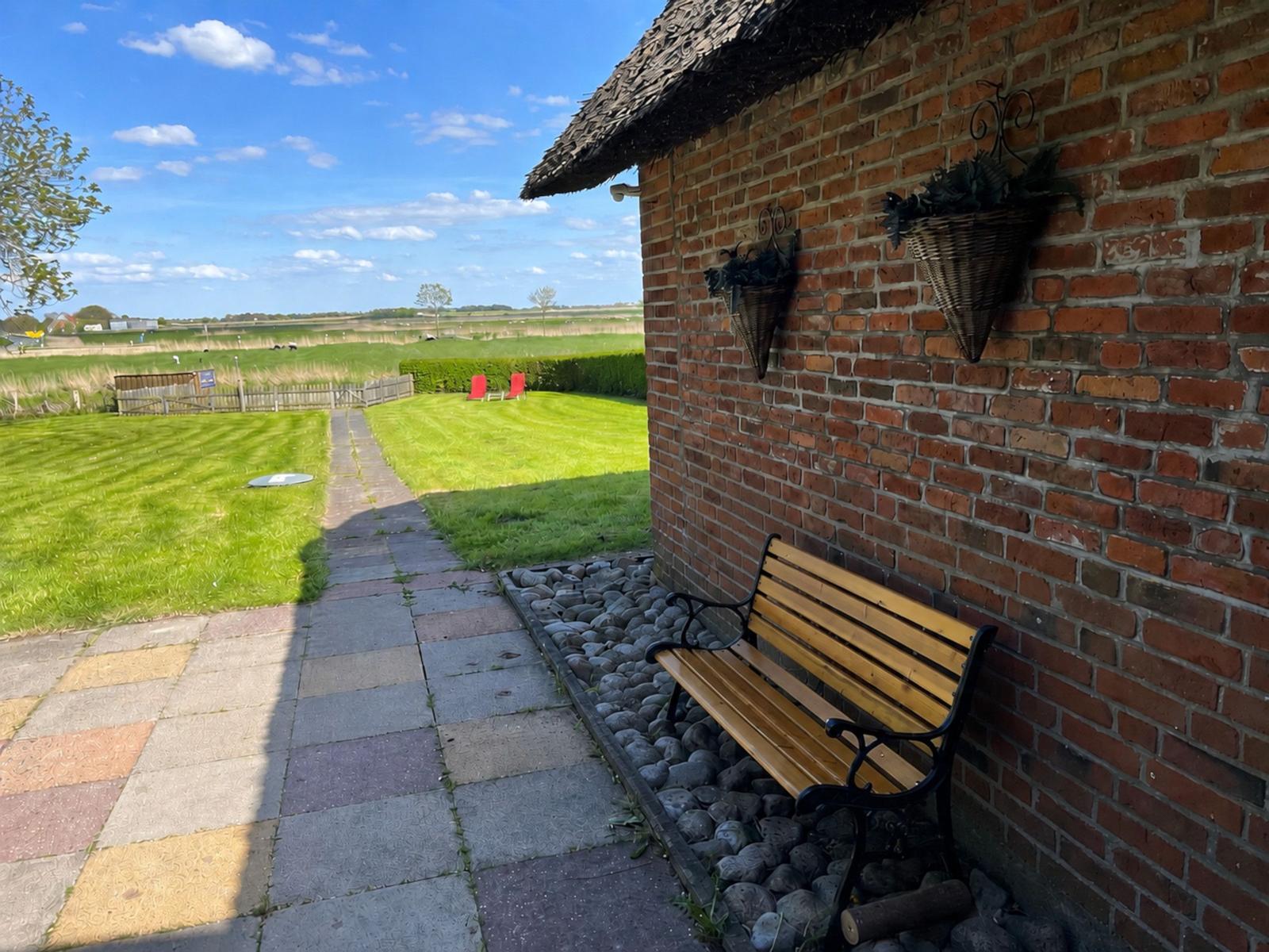 Holzbank vor Backsteinmauer mit Steinweg und grünem Rasen im Hintergrund.