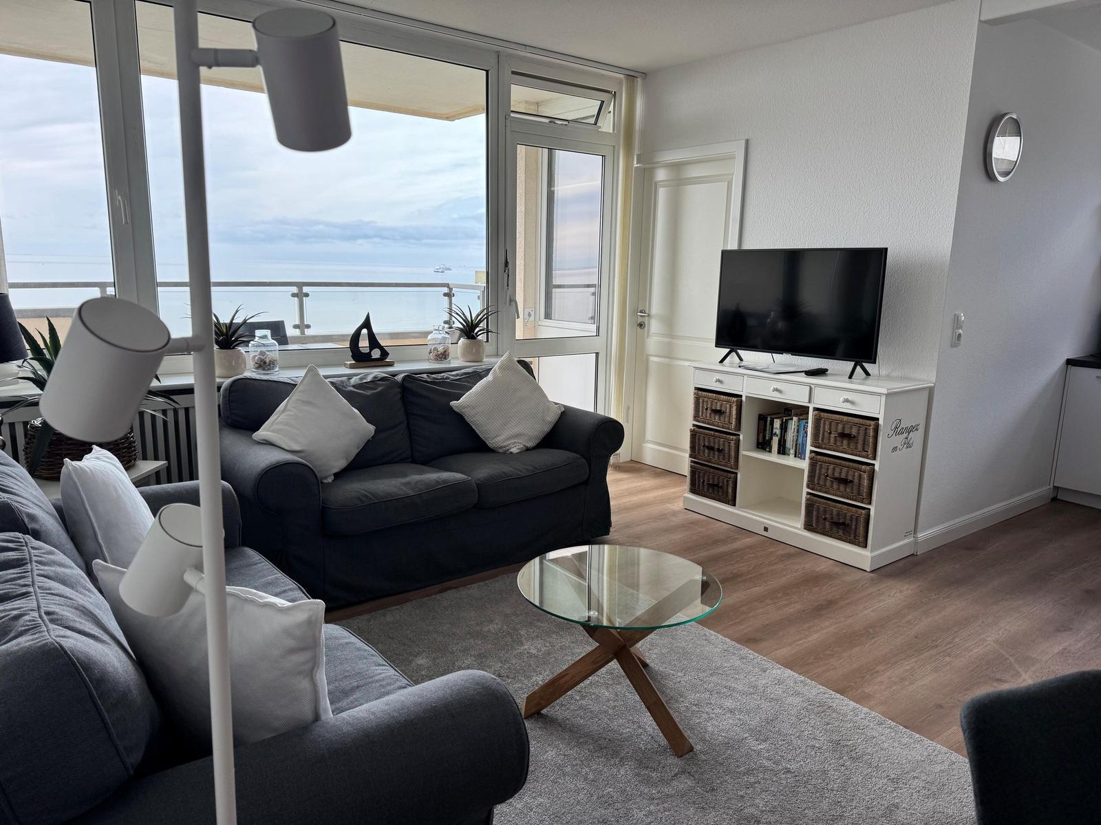 Wohnzimmer mit Sofa, Fernseher und Meerblick durch große Fenster.