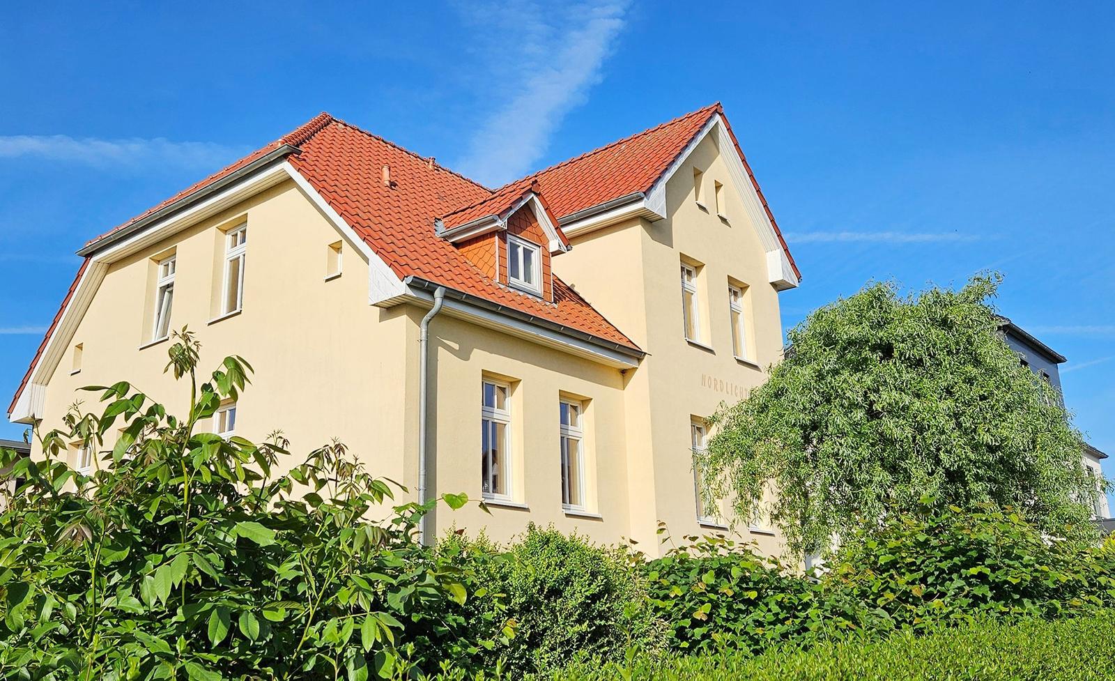 Ein zweistöckiges Haus mit rotem Dach und hellen Wänden, umgeben von Grünflächen.