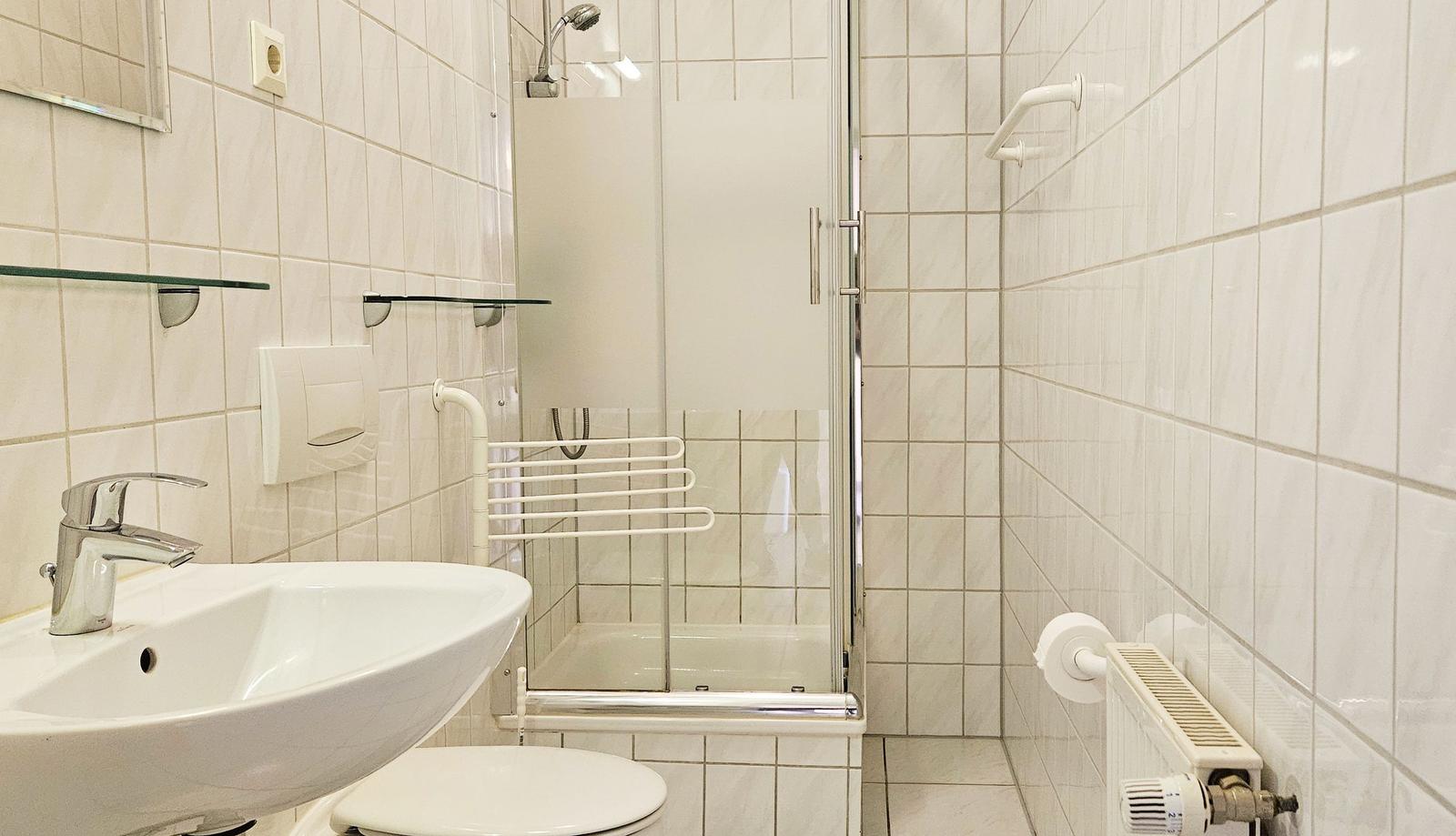 Badezimmer mit Dusche, Waschbecken und Toilette. Weiße Fliesen und Handlauf.