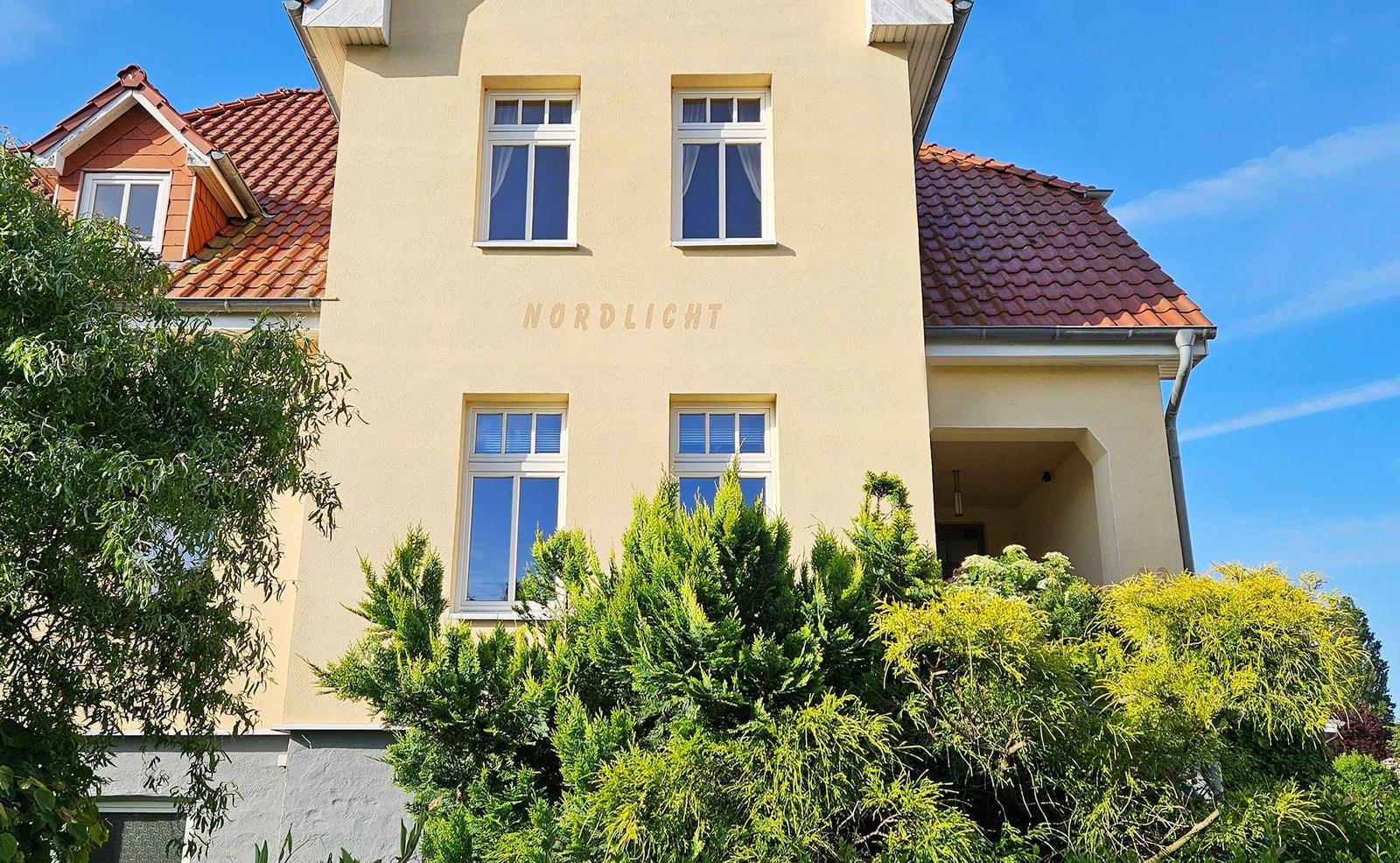 Haus mit gelbem Putz, rotem Dach und grüner Vegetation. Auf der Fassade steht NORDLICHT.