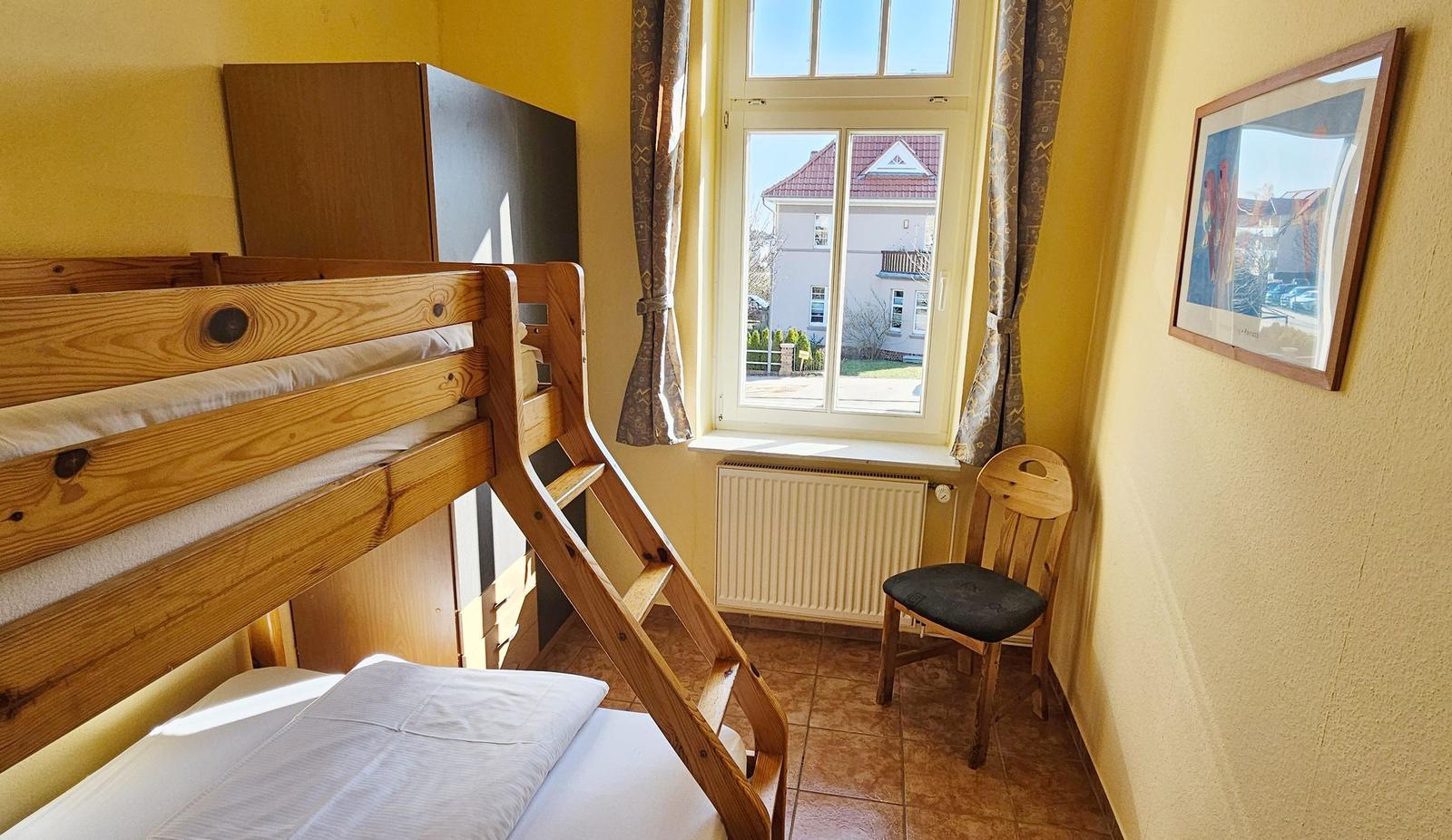 Zimmer mit Holzbett, Fenster und Stuhl.