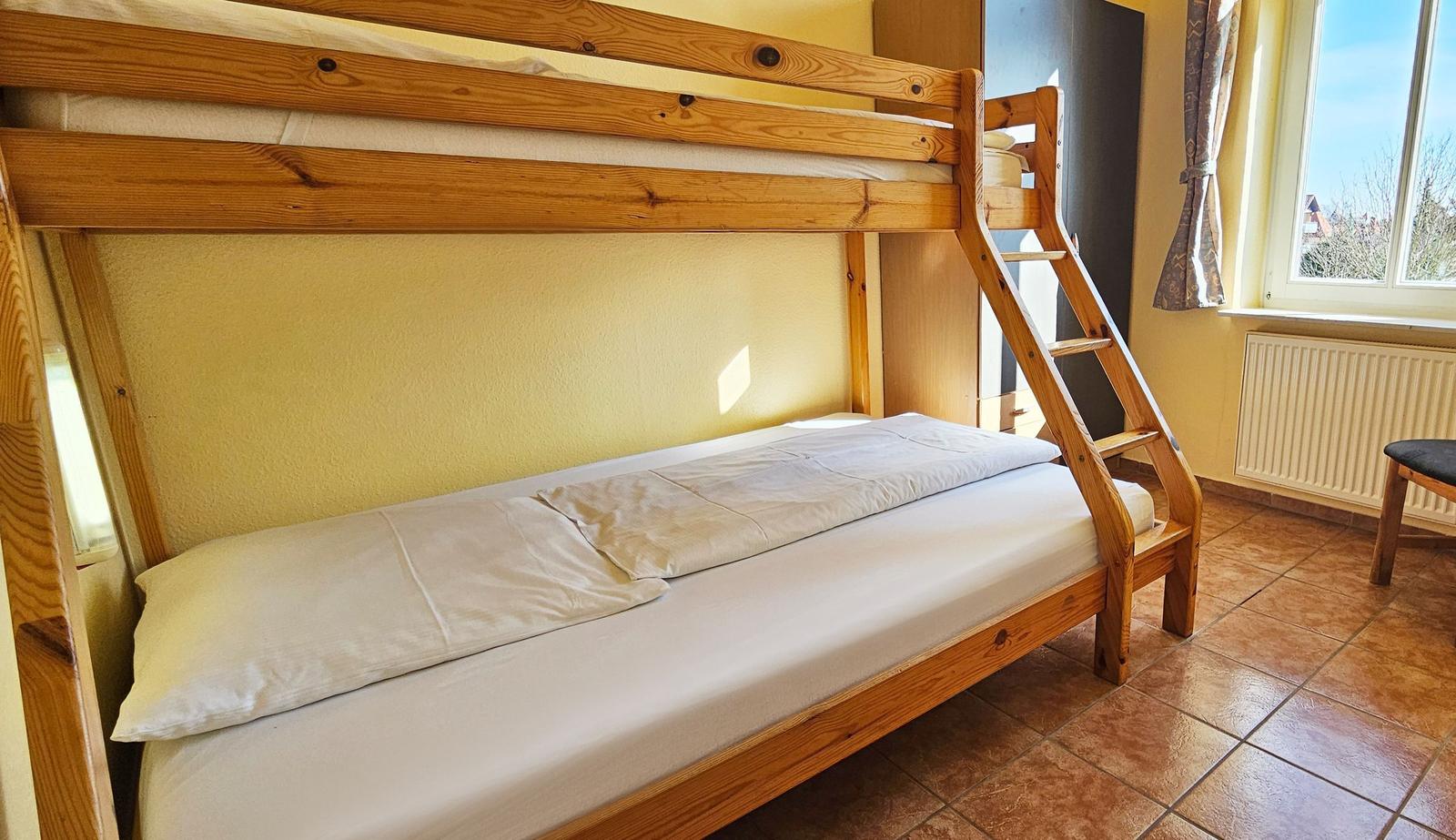 Ein Doppelstockbett mit Holzgestell und weißer Bettwäsche in einem Raum mit Fenster und Heizkörper.