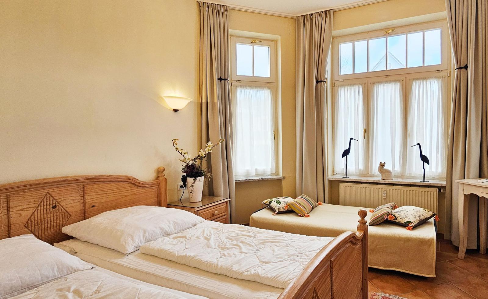 Zimmer mit zwei Betten, Fenstern und Vorhängen. Auf dem Fensterbrett stehen zwei Vogelfiguren und eine Katze.