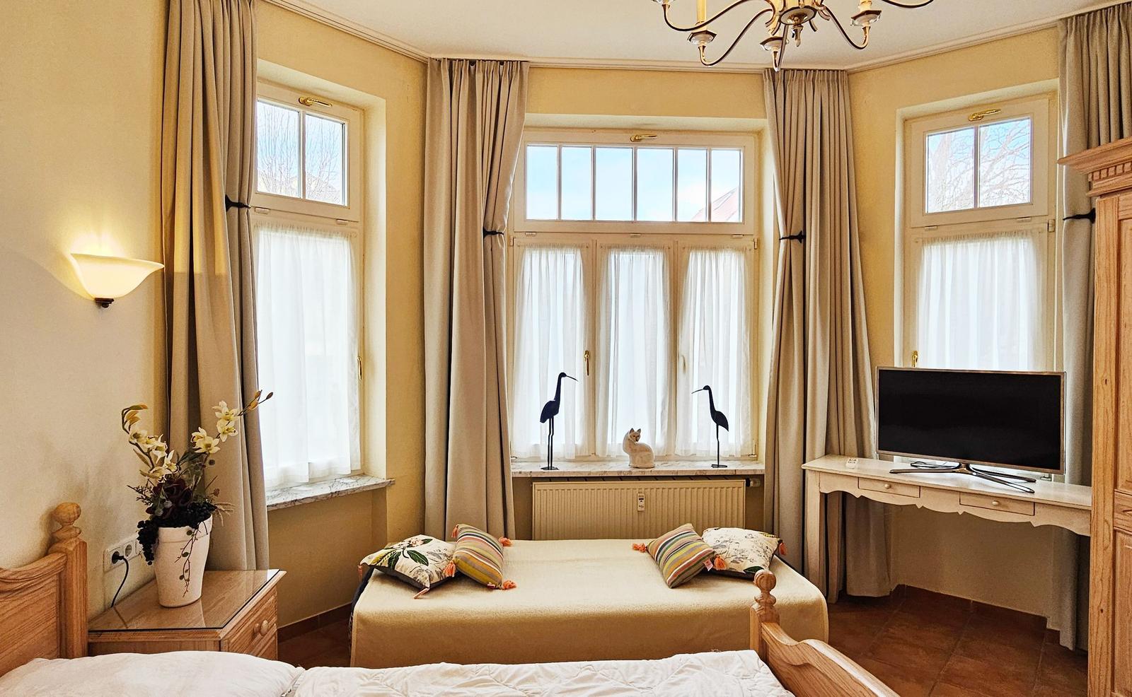 Schlafzimmer mit Bett, TV, drei Fenstern und Holzmöbeln.