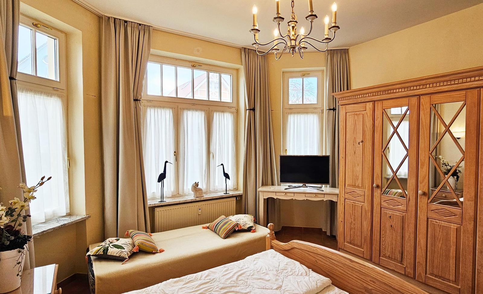 Schlafzimmer mit Doppelbett, Holzschrank, TV und Fenstern mit Vorhängen.