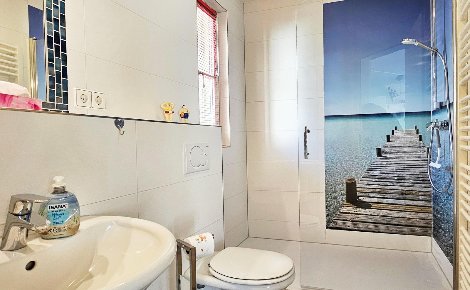 Bad mit Dusche, Waschbecken und Toilette. Duschwand zeigt Strandmotiv.