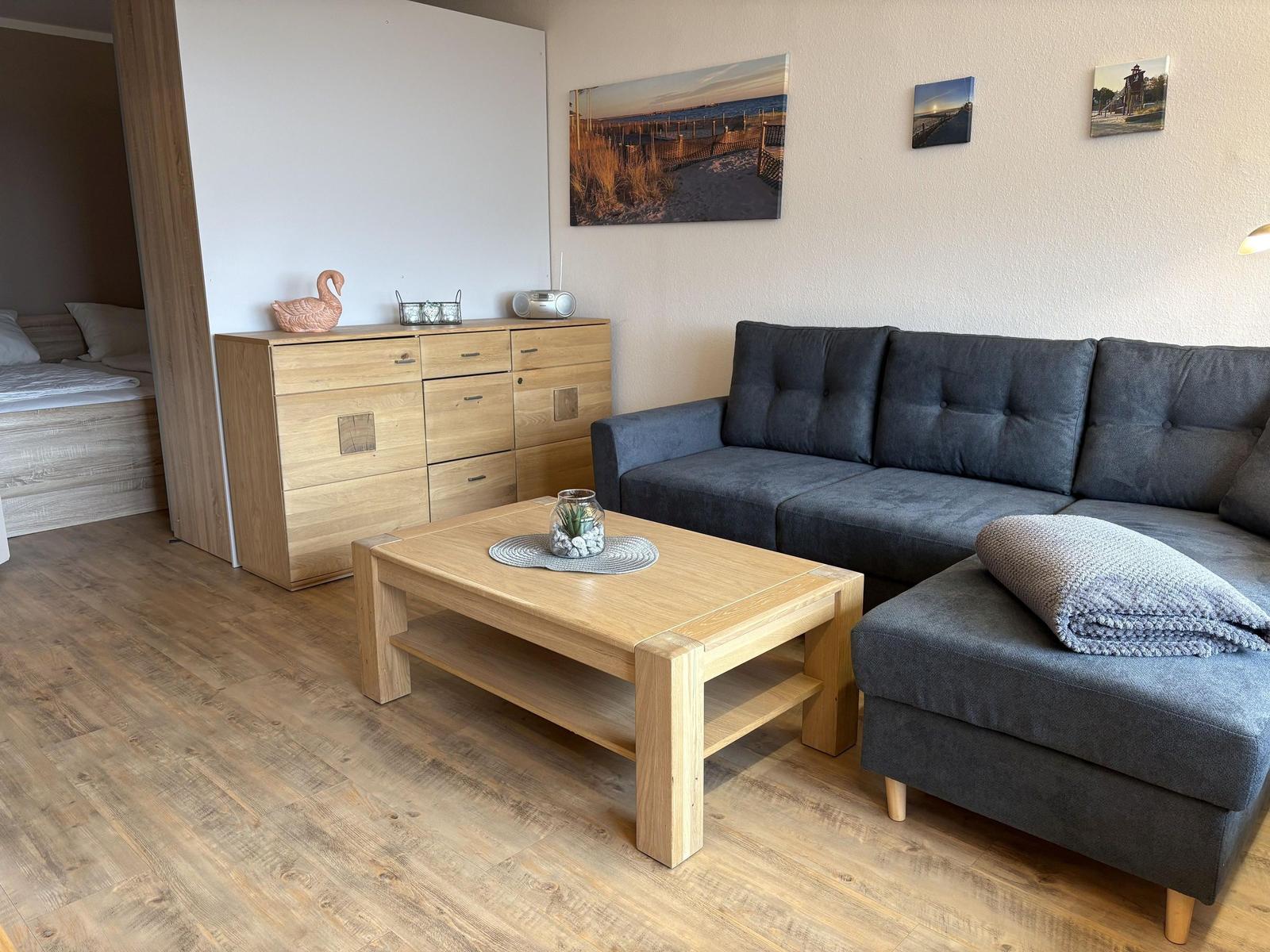 Ein Wohnzimmer mit grauem Sofa, Holztisch und Schrank. Wandbilder zeigen Strand.