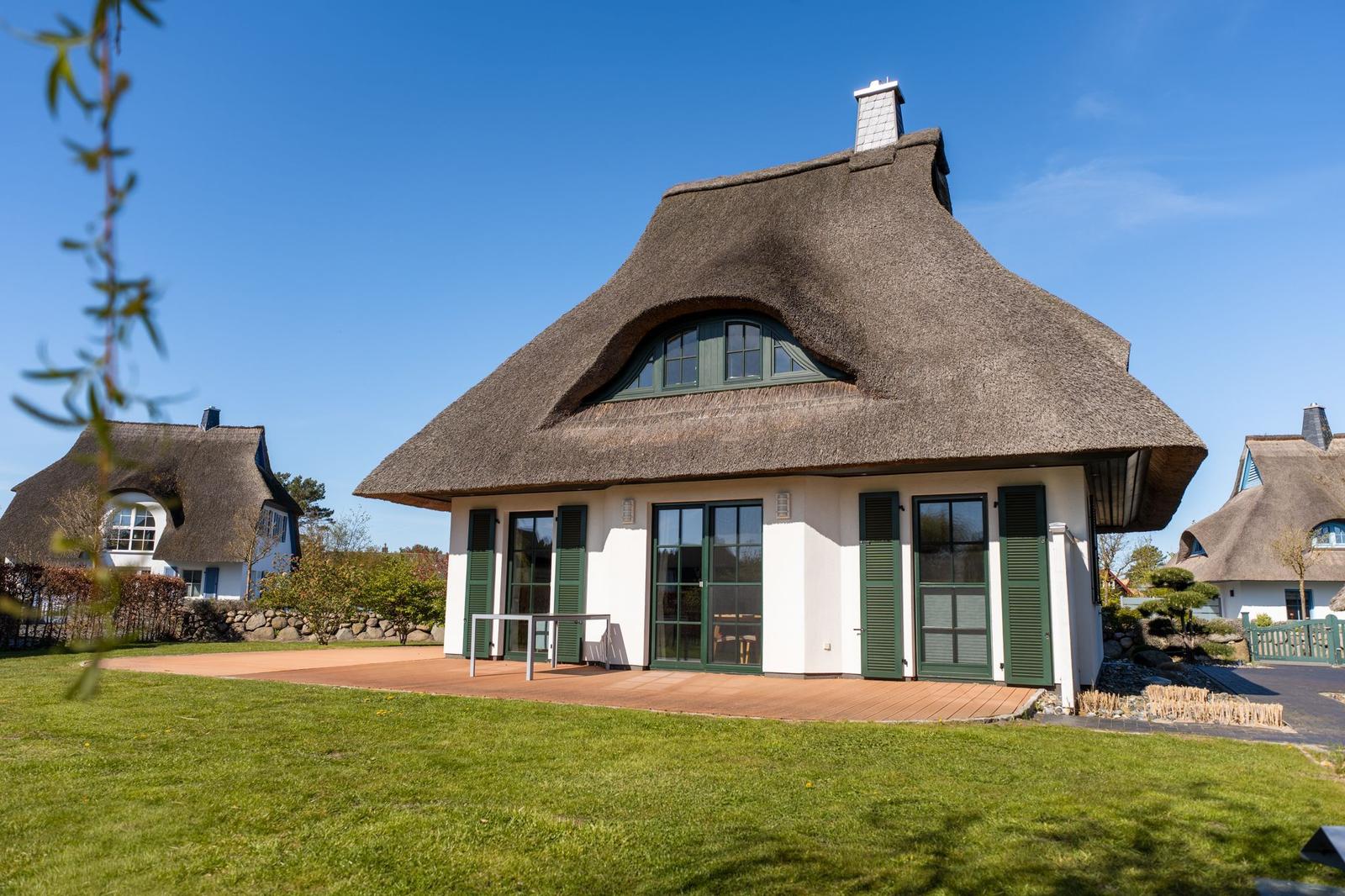 Haus mit Strohdach, grünen Fensterläden und Terrasse auf grünem Rasen.