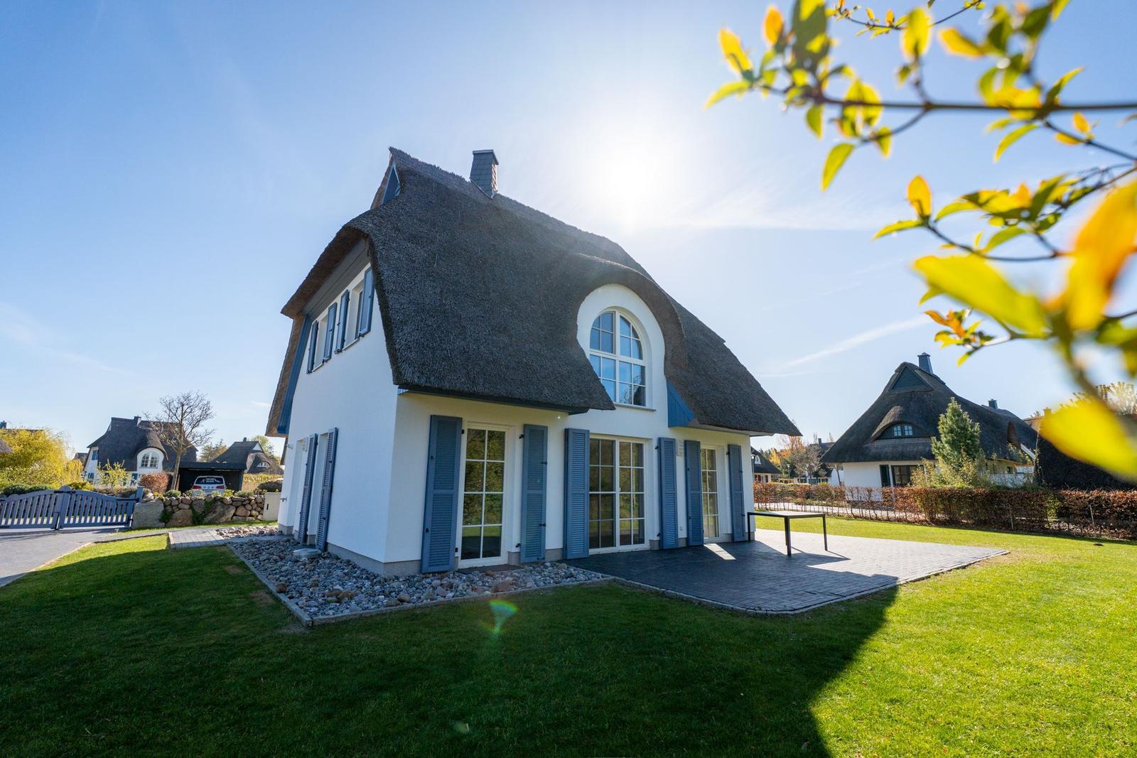 Ein weißes Haus mit strohgedecktem Dach und blauen Fensterläden steht auf einer grünen Wiese.