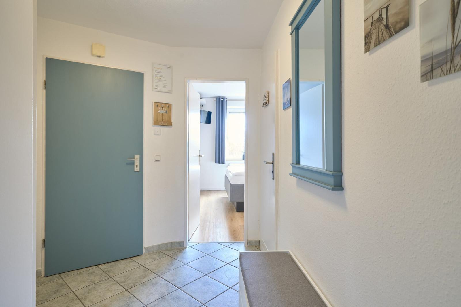 Flur mit blauer Tür, Spiegel und Bildern an der Wand. Durchgang zu einem Schlafzimmer mit Bett.