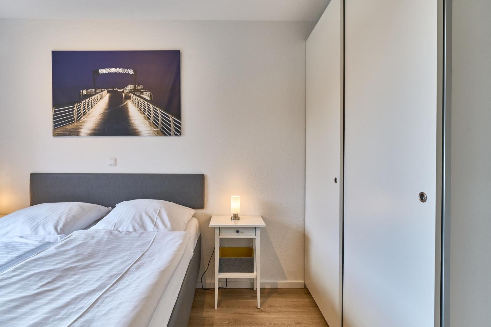 Schlafzimmer mit Bett, Nachttisch und Wandbild. Kleiner Schrank an der Wand.