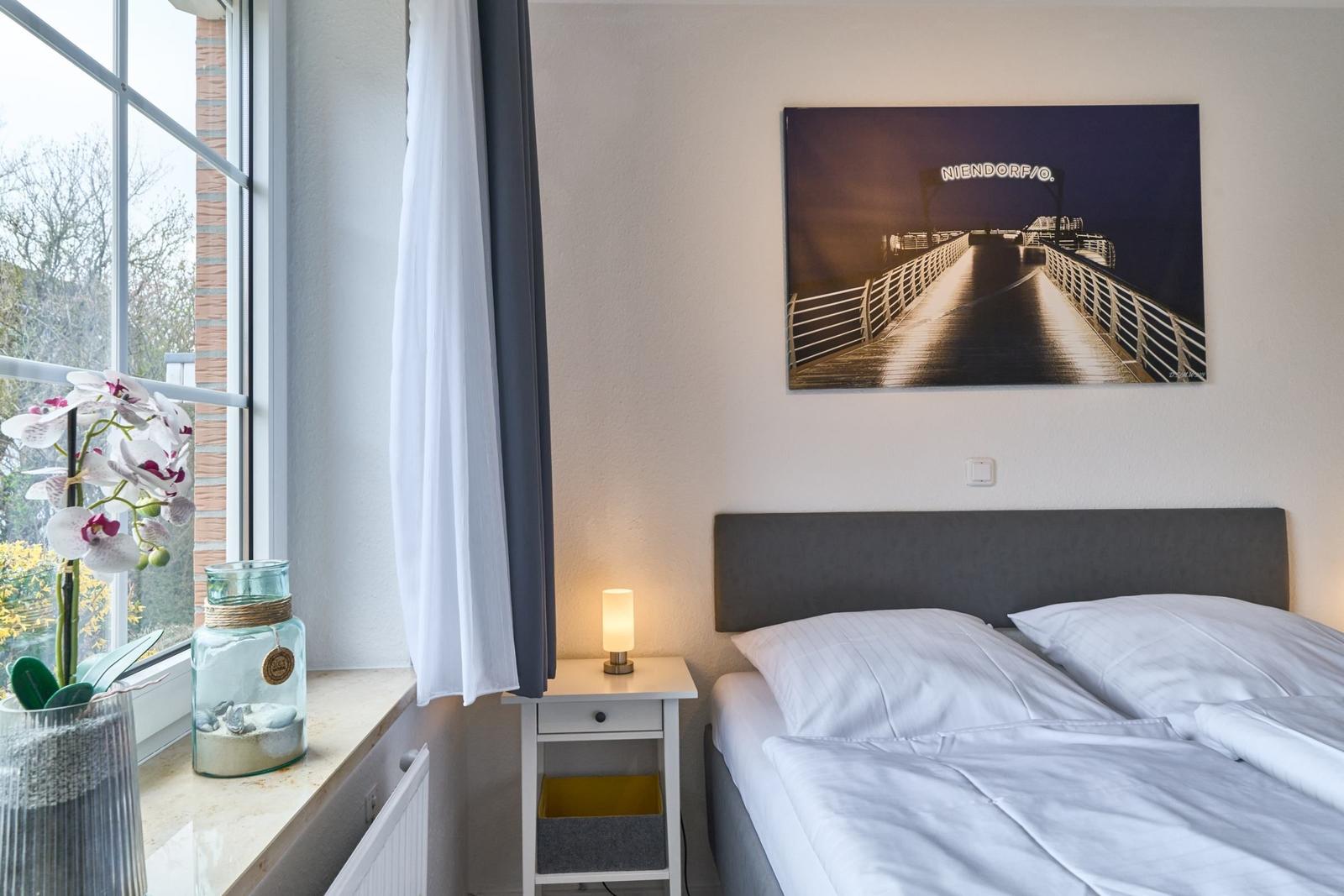Schlafzimmer mit Bett, Nachttisch und Fenster. Wandbild zeigt Brücke mit Schriftzug.