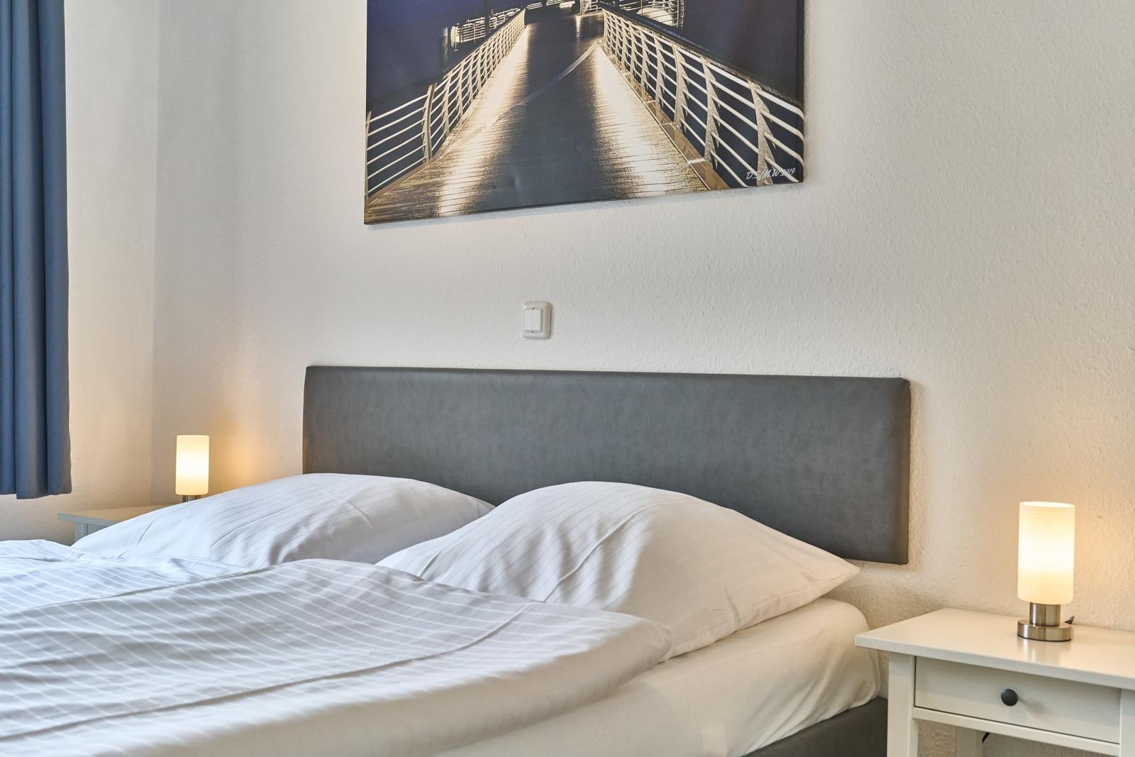 Doppelbett mit weißen Kissen und Bettwäsche, zwei Nachttische mit Lampen, Wandbild über dem Kopfteil.